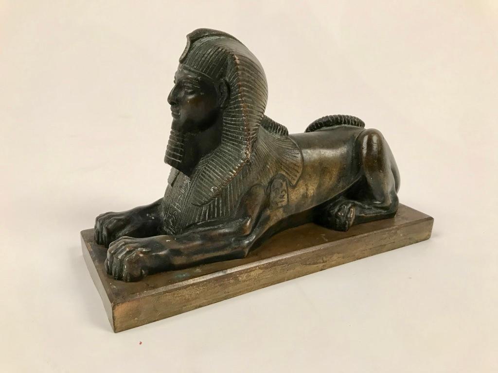 A French grand tour bronze model of a reclining sphinx, early 19th century. The recumbent sphinx shown with elaborate headdress, its tail curled onto its back, seated on a rectangular base.

The use of sphinxes as a motif for bronze objects