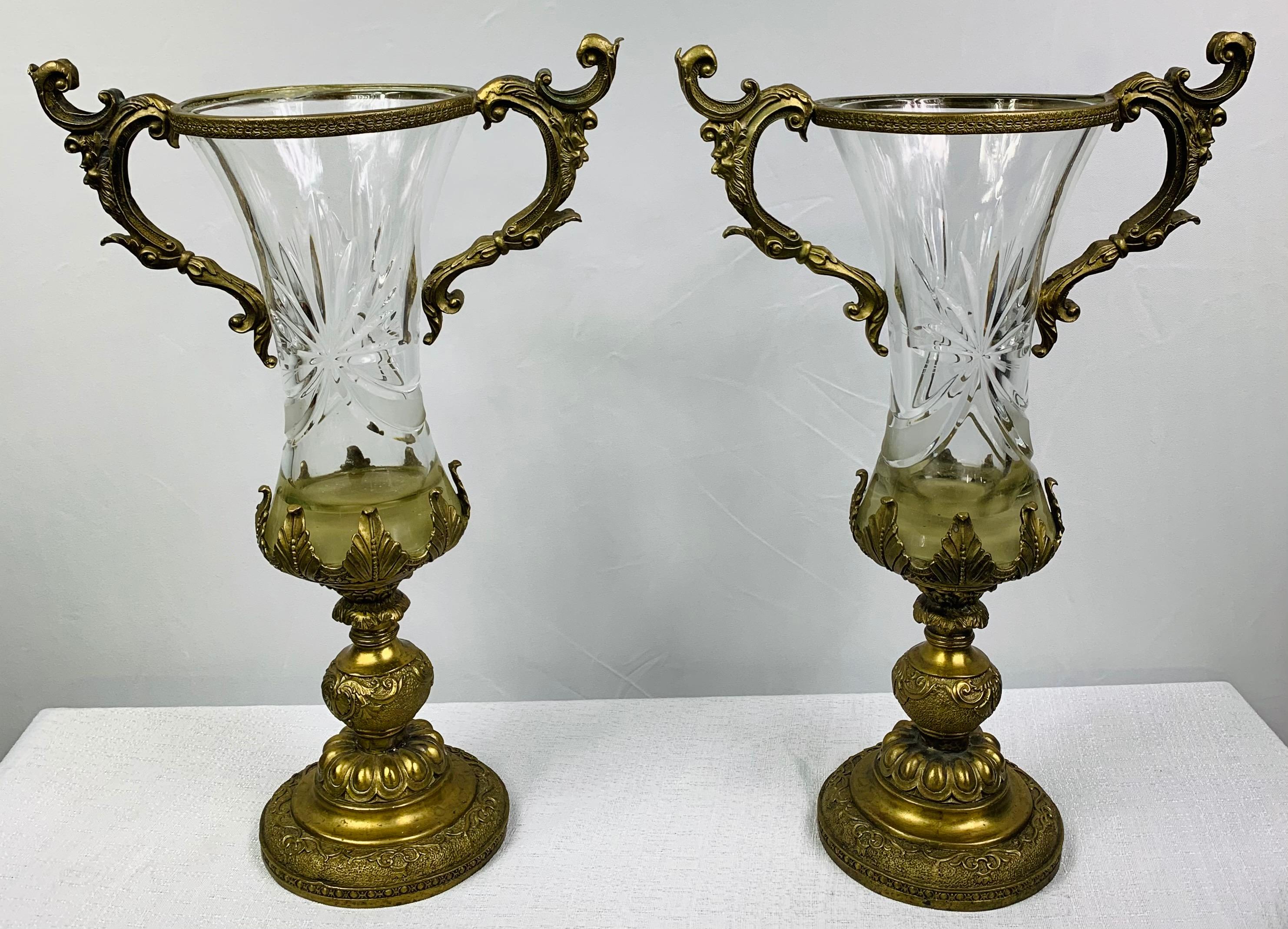 An exquisite pair of early 20th century bronze mounted and cut glass twin handle vases or urn in the manner of Baccarat. Each vase bodies are crafted of cut-glass and feature a round base. The handles, base and mouth is finely decorated in bronze