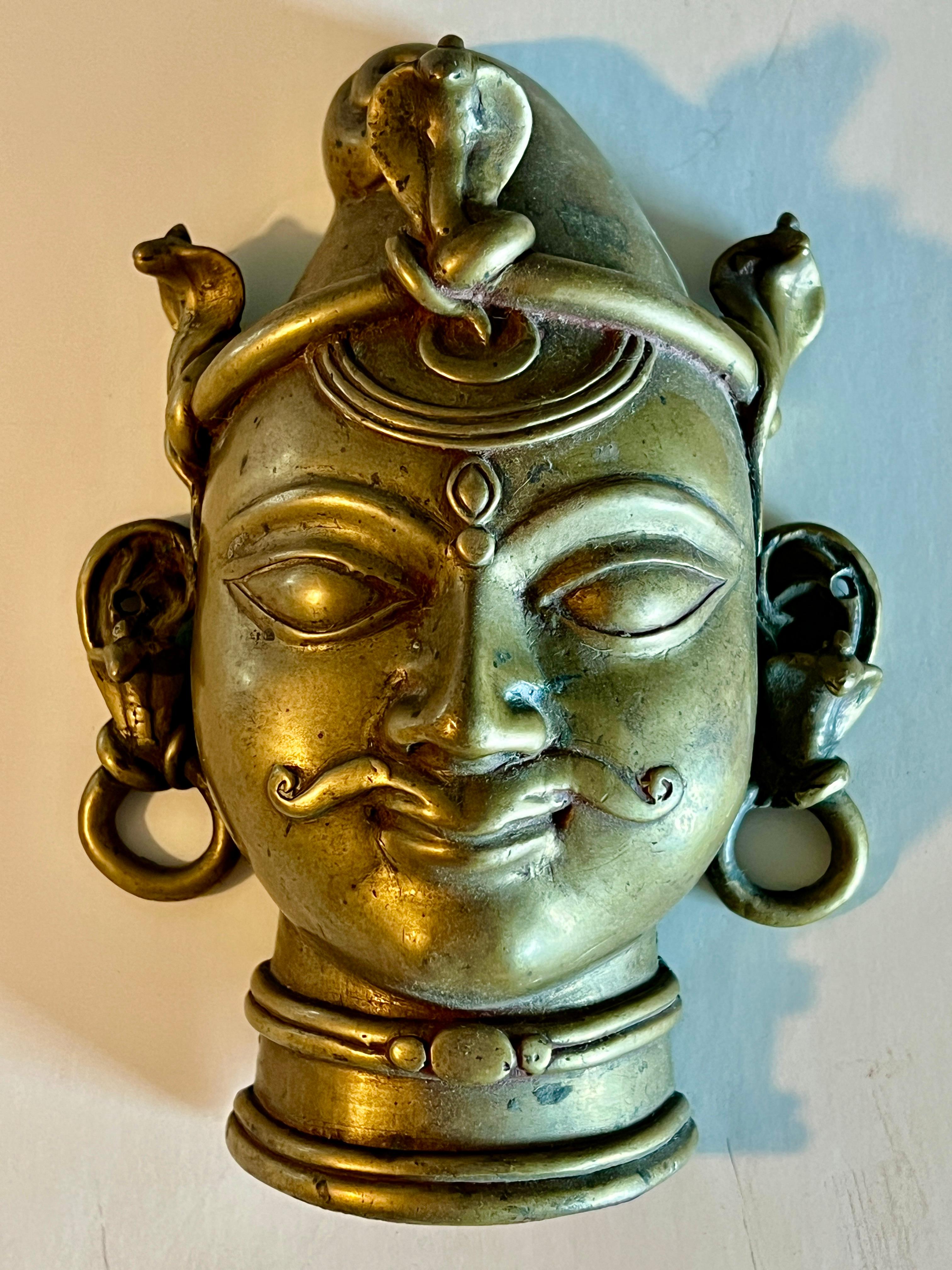 Questa placca di bronzo raffigurante Shiva con un volto umano era destinata a fungere da copertura del lingam.  Questi esempi hanno dei baffi elaborati,  un cappuccio orlato di cobra arrotolati, occhi a mandorla, orecchini, una collana, una luna e