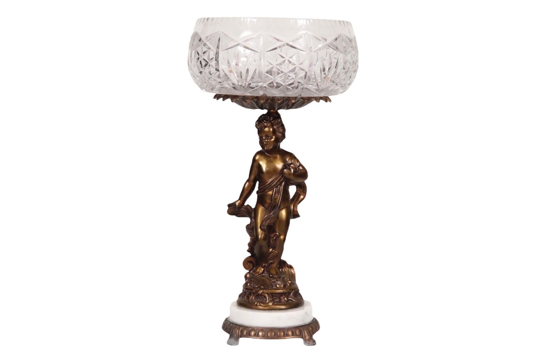 A large figural centerpiece compotier. A cut glass bowl with a sawtooth edged rim and faceted pattern is supported by a bronze plated cast cherub. On the cherub's head is a bronze bobeche in the shape of fanned feathers. The base consists of round
