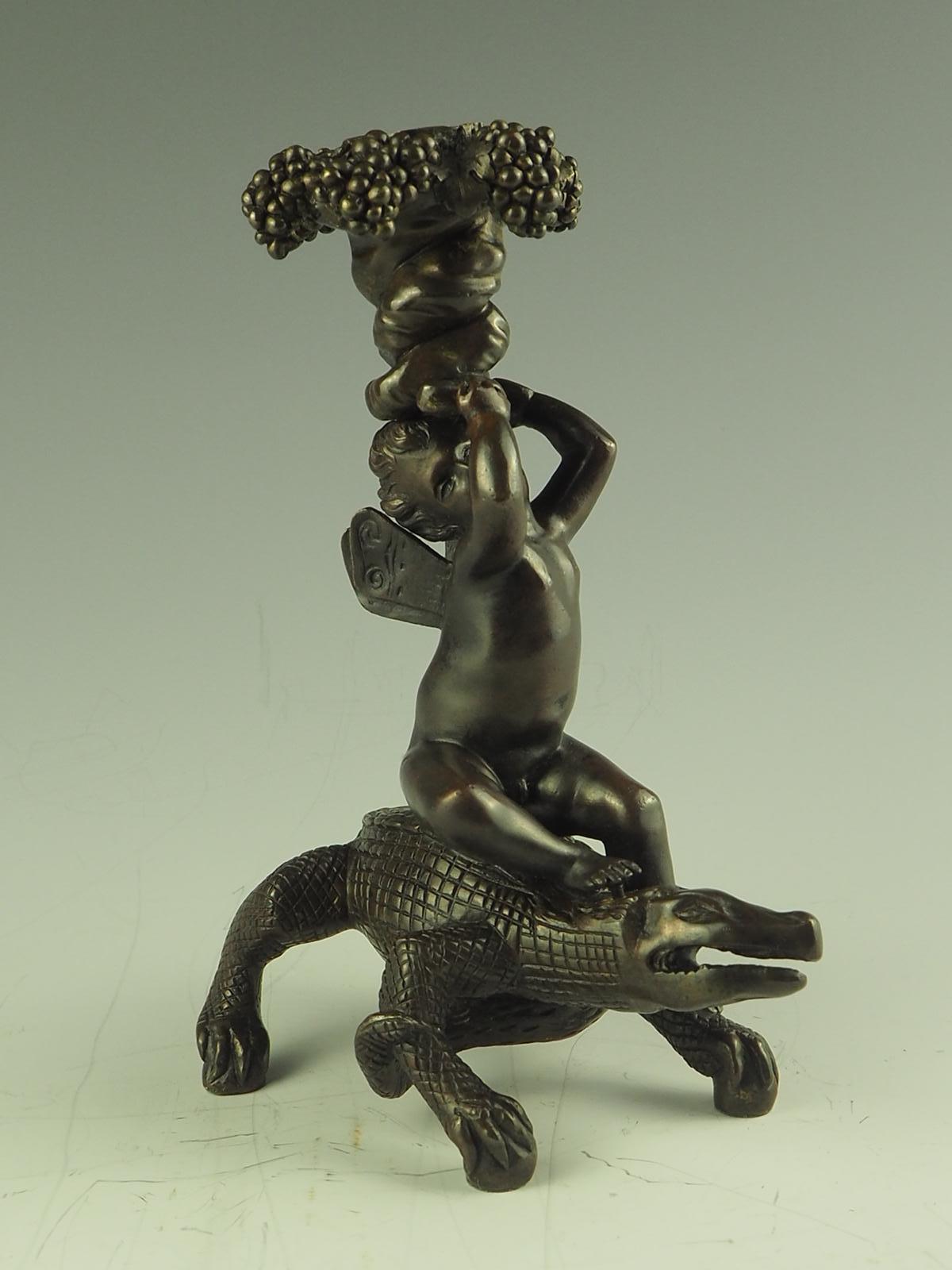 Antique bronze candlestick, depict a bacchanalian putto astride crocodile (or alligator), holding aloft entwined vines, rising to bunches of grapes and vine leaves. The putto is sat on a crocodile, with mouth open and tail twist beneath.

c 1890.