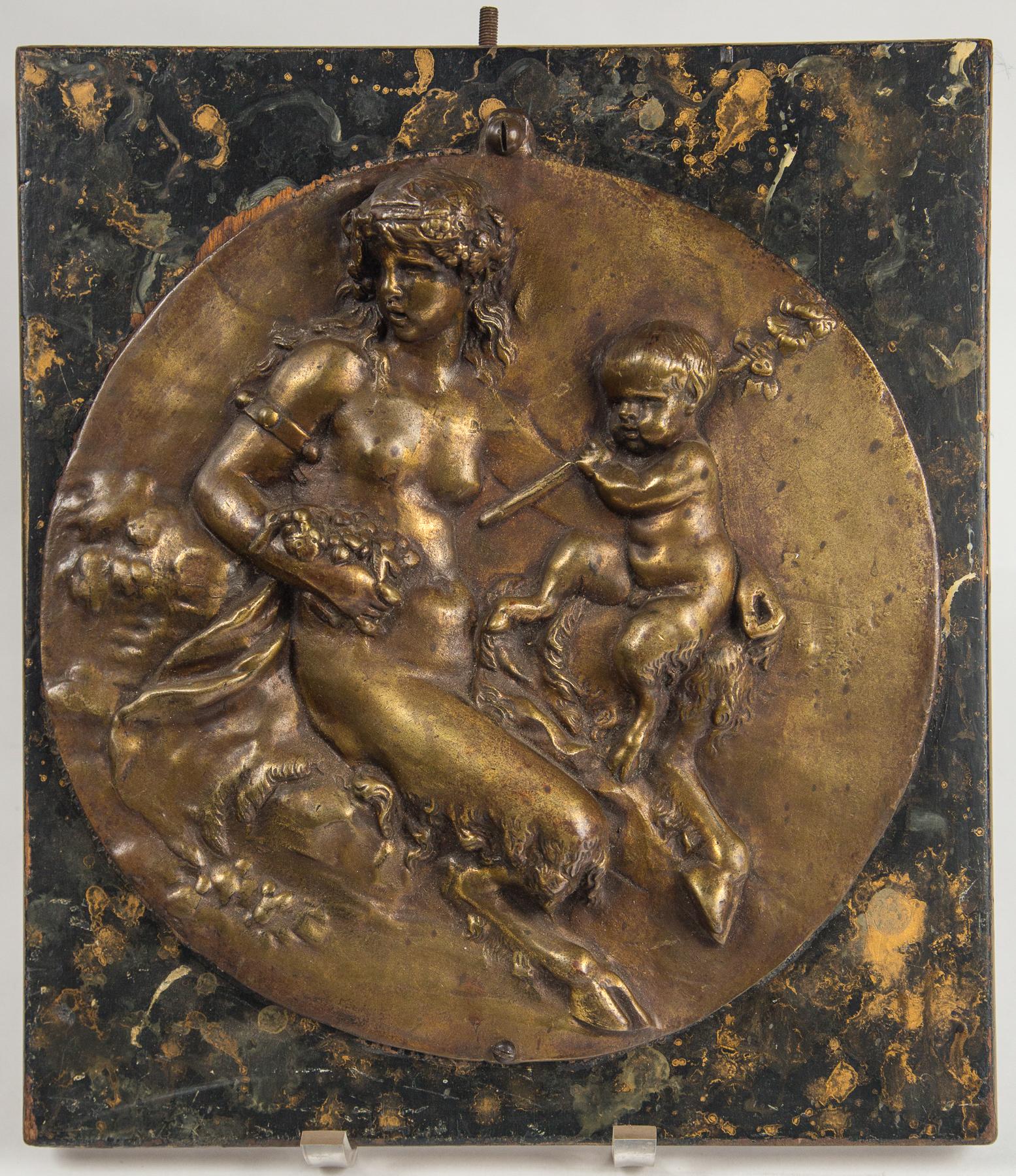 The plaque is attached, with screws, top and bottom, to a faux marble painted piece of wood measuring 13.5 x 12 x 1. This plaque depicts a female and child satyr. They each have hoofed feet. It seems to be finished in dark gold tone lacquer on the