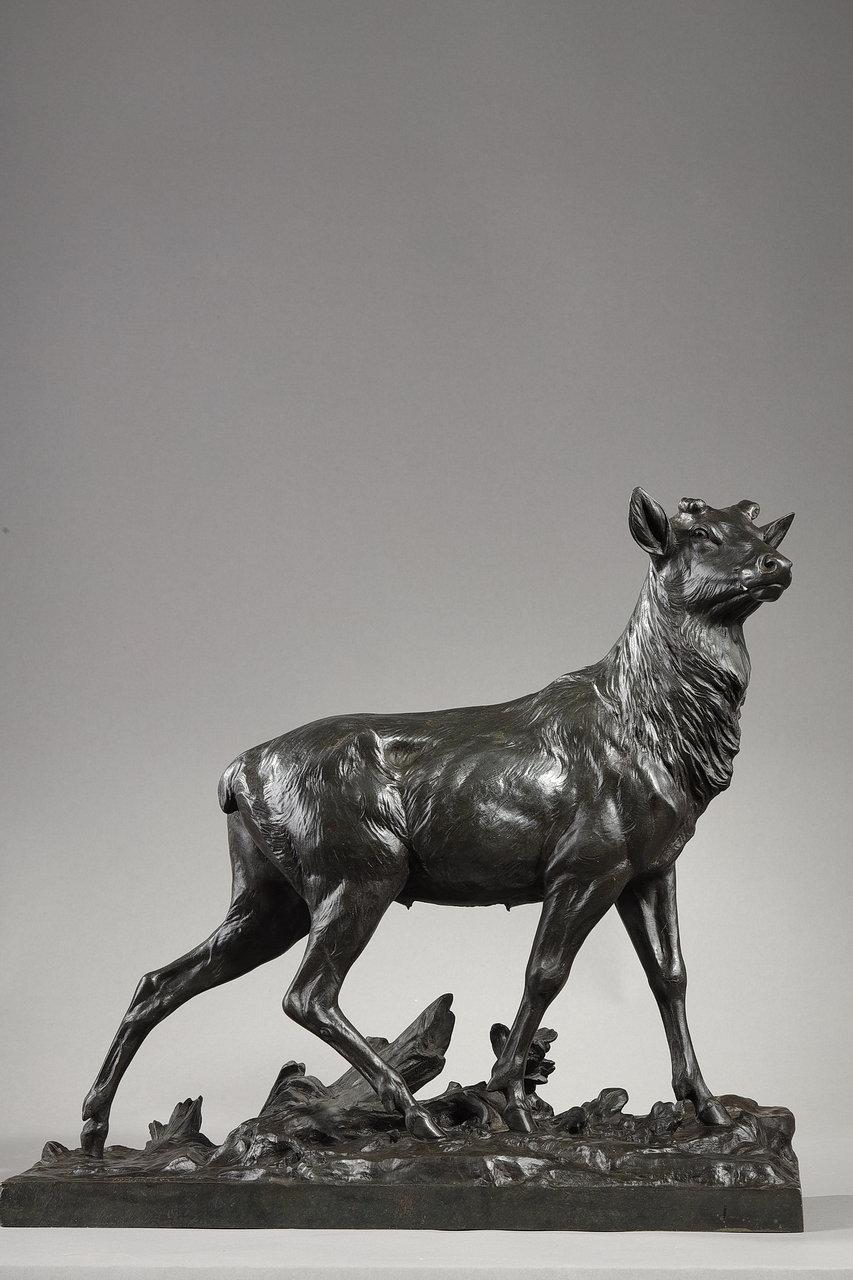 Skulptur aus Bronze mit brauner Patina, die einen Hirsch nach der Mauser darstellt. Er ruht auf einer Terrasse, die einen Sockel bildet, der von Baumstümpfen bewegt wird und mit Eichenlaub und Eicheln übersät ist. Das Fell des Tieres ist realistisch