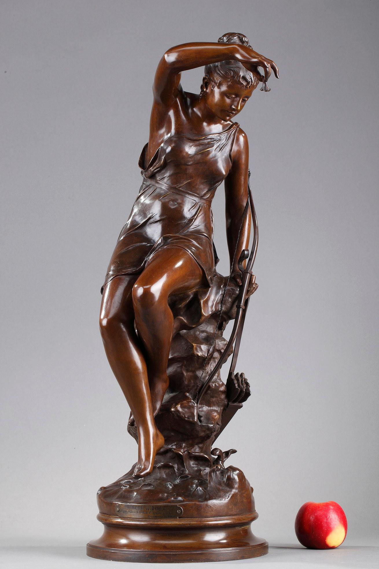 Belle statue en bronze patiné représentant Diane, déesse de la chasse, également appelée Nymphe de Diane, d'après l'œuvre de Lucie Signoret-Ledieu. Elle représente la déesse assise sur un tronc tenant son arc dans une main et de l'autre une fleur en