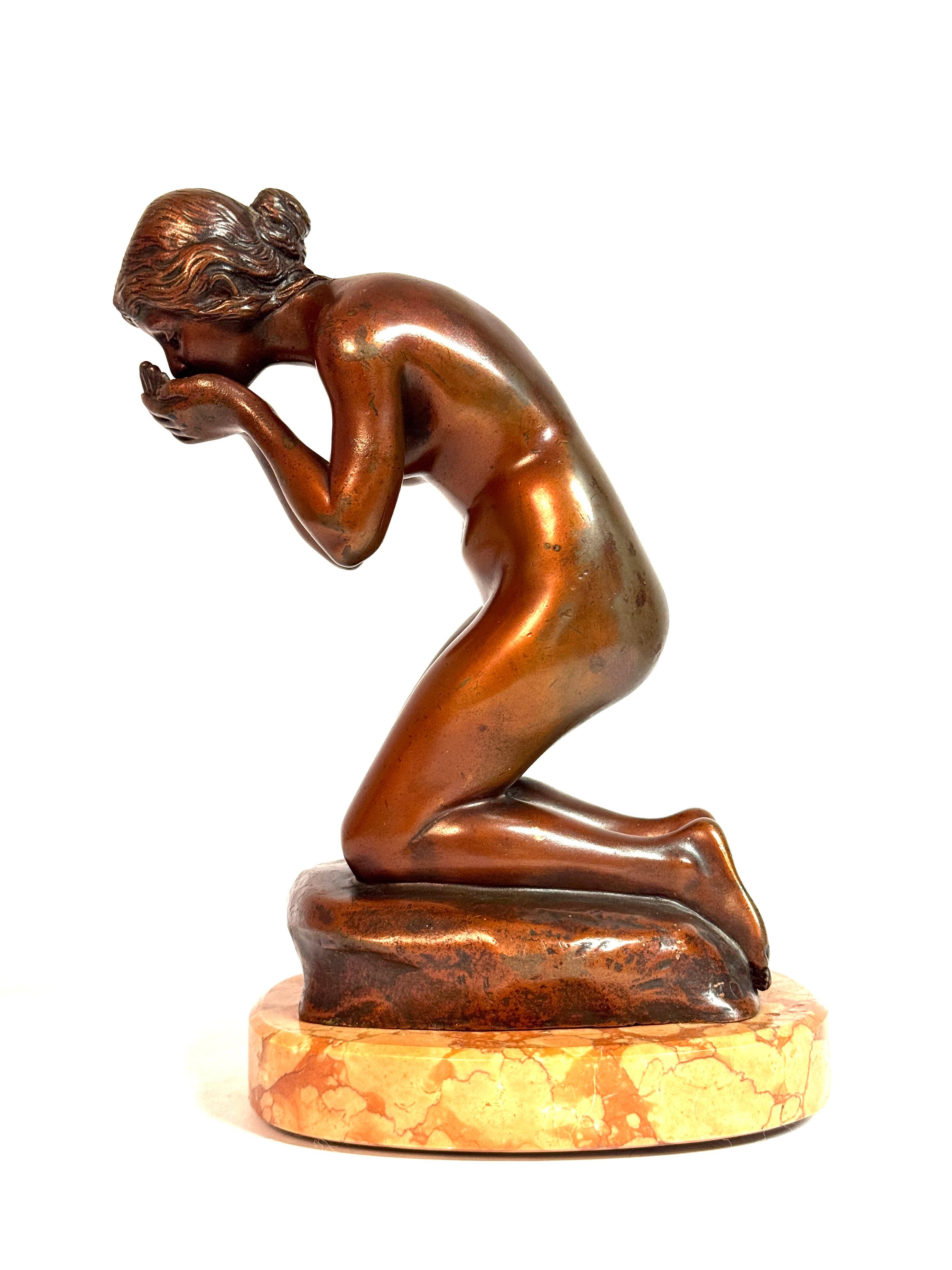 Questa squisita scultura in bronzo dello scultore tedesco Rudolf Kaesbach (1873-1955) raffigura una giovane donna inginocchiata su una base naturalistica, mentre si stringe delicatamente le mani per bere. La figura è catturata in una posa intima e