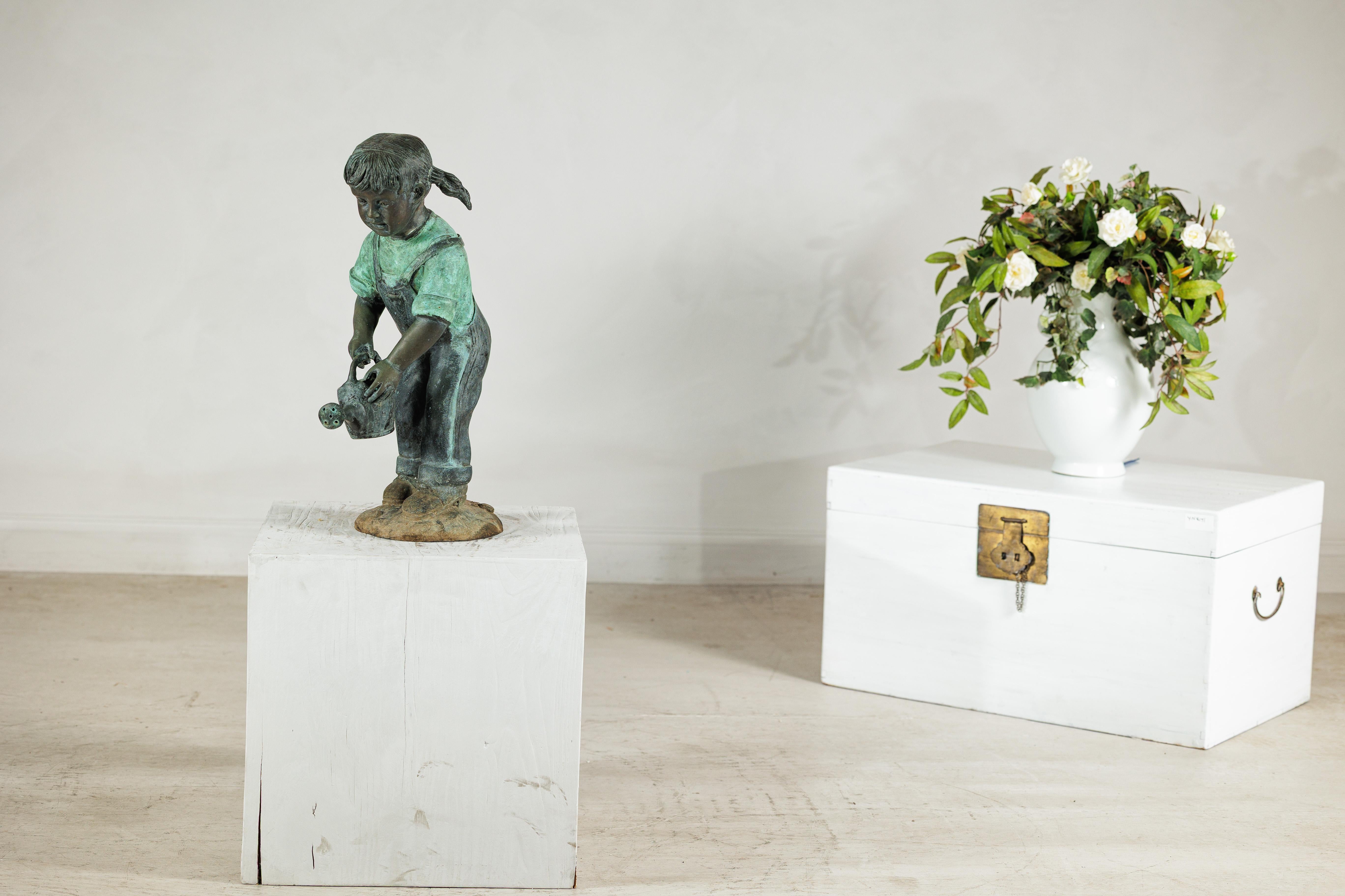 A bronze garden sculpture depicting a young girl in pigtails, dressed in rolled overalls and a short-sleeved shirt, leaning forward as she pours from a watering can. The figure is tubed for fountain use, designed so that water may flow naturally