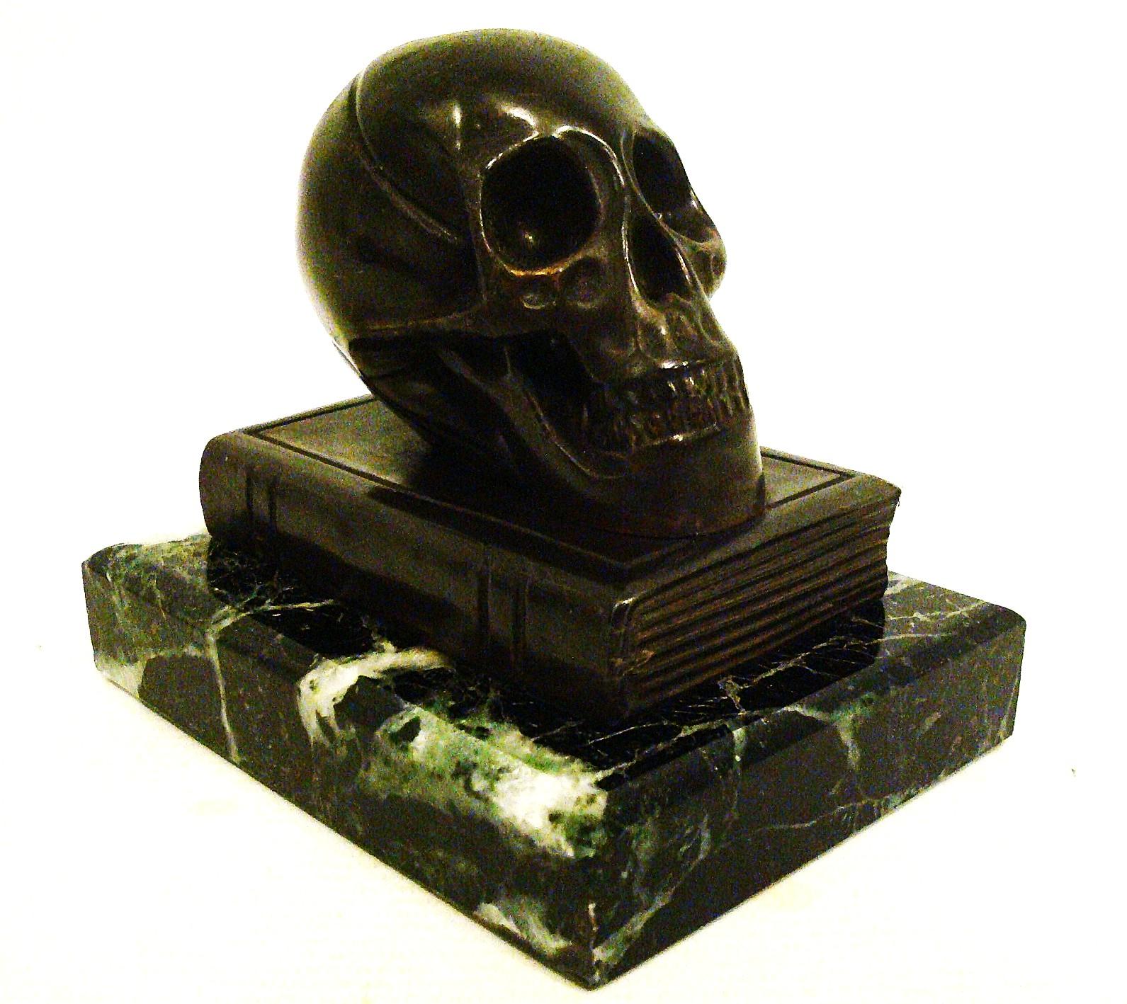 Escultura / Pisapapeles de bronce con forma de calavera sobre un libro.
Esta escultura es un trueno silencioso: elegante, solemne y rico en peso filosófico. La calavera sobre el libro cerrado, apoyada en un pedestal jaspeado, parece una vanitas