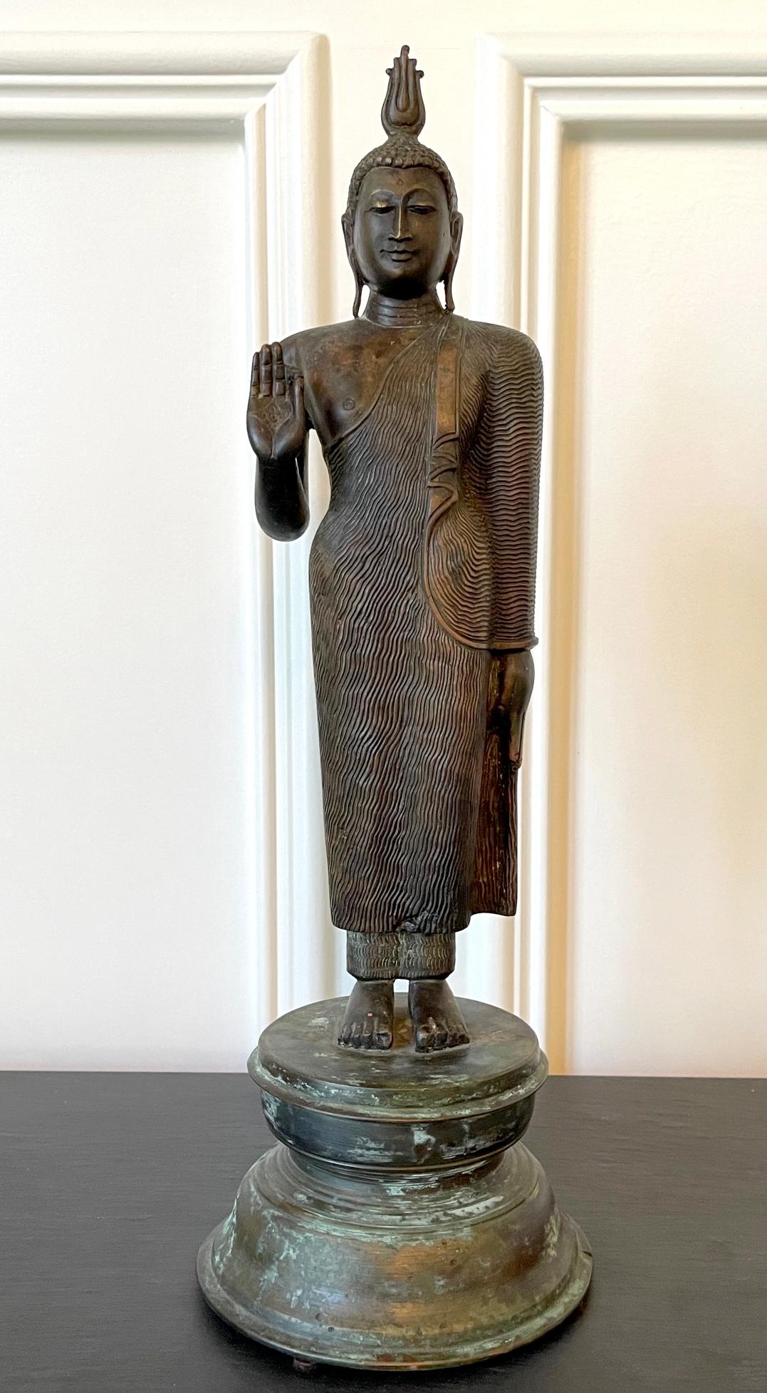 A standing Buddha statue of cast bronze from southeast Asian island nation Sir Lanka circa late 19th to early 20th century. The upright statue is of a straight frontal stance with his right hand raised in Abhaya mudra, a symbol to dispel fear and