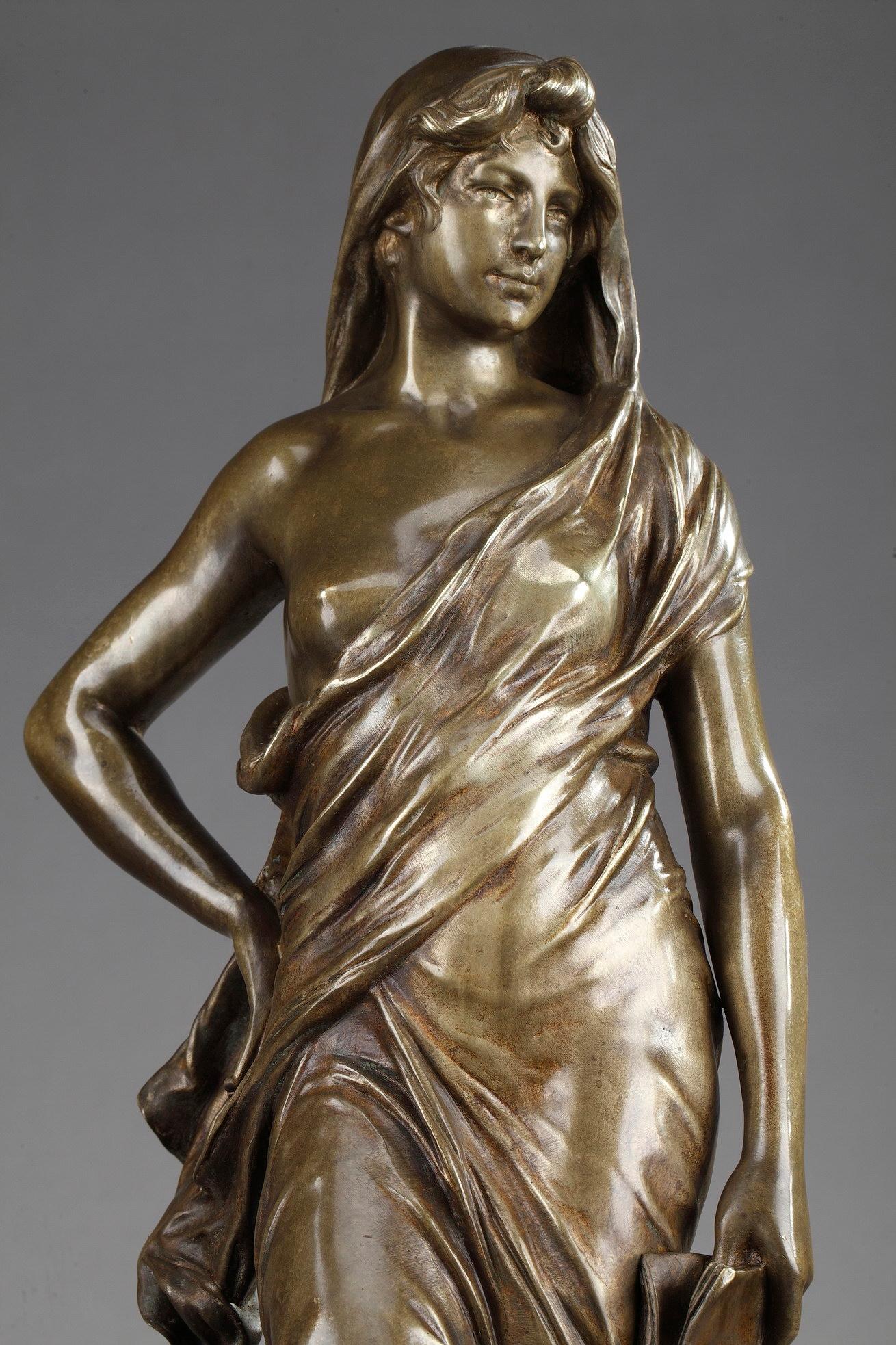 Bronze statue with gold patina featuring an allegory of Meditation. The young woman holds a book in one hand, while the other gently rests upon her hip. She is wearing a flowing dress which covers partially her head and breast. This sculpture boasts