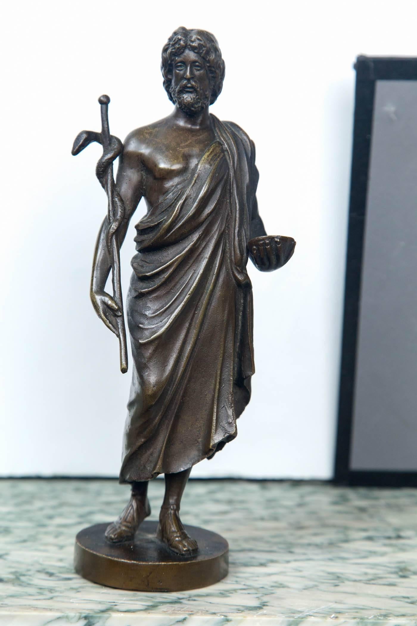 Standing upon a base, the patinated statue holds a staff cradled in his right hand and arm, with a snake wrapped around it. (the cadeusus). In his left hand he holds a bowl. He wears a toga

On the back side of the base is the name Benetti. We