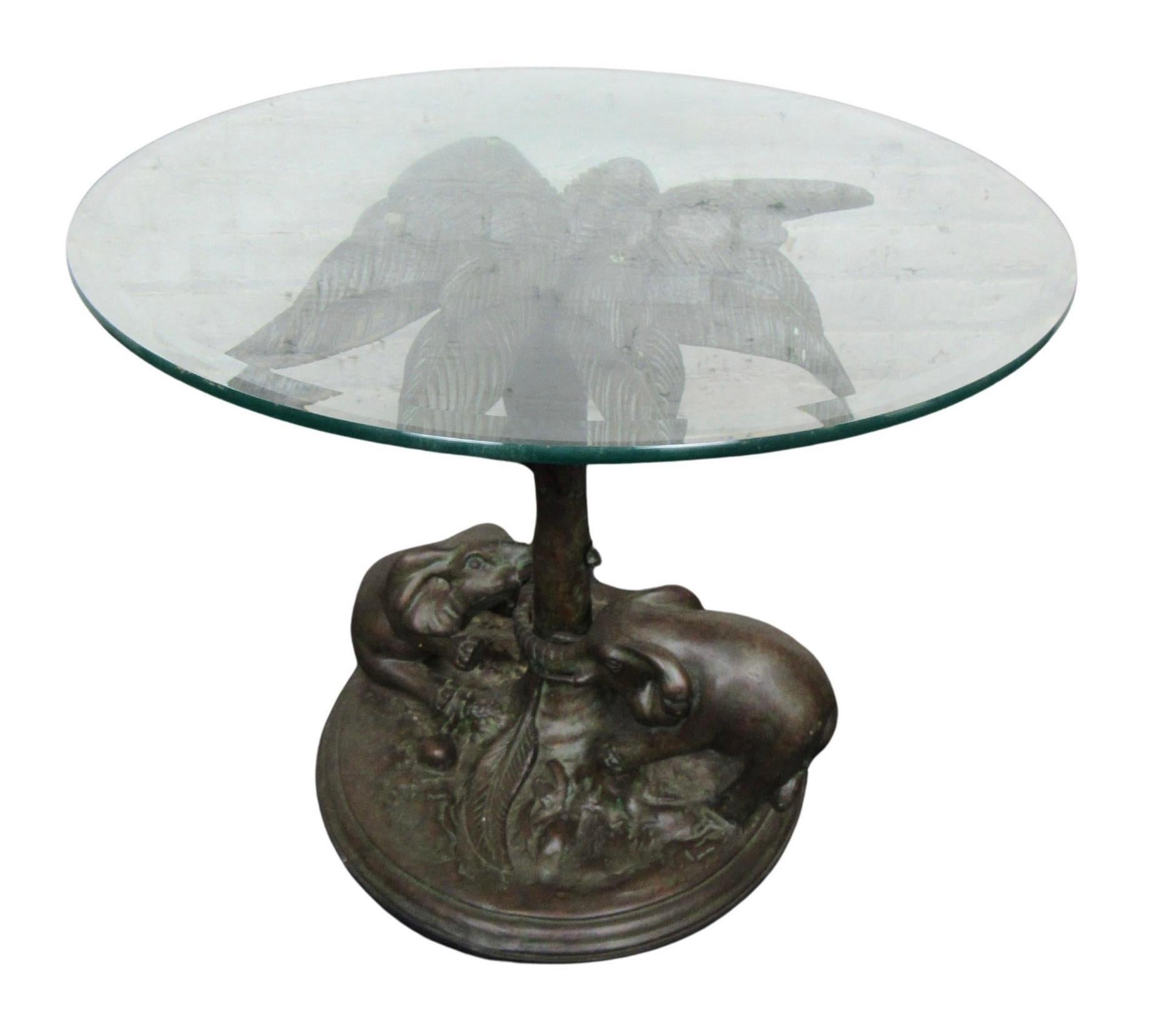 Esta mesa auxiliar tiene un pesado tablero de cristal redondeado que descansa sobre una elaborada base esculpida de color bronce. La base utiliza una palmera para sostener la parte superior de la mesa, y dos pequeñas figuras de elefantes adornan la