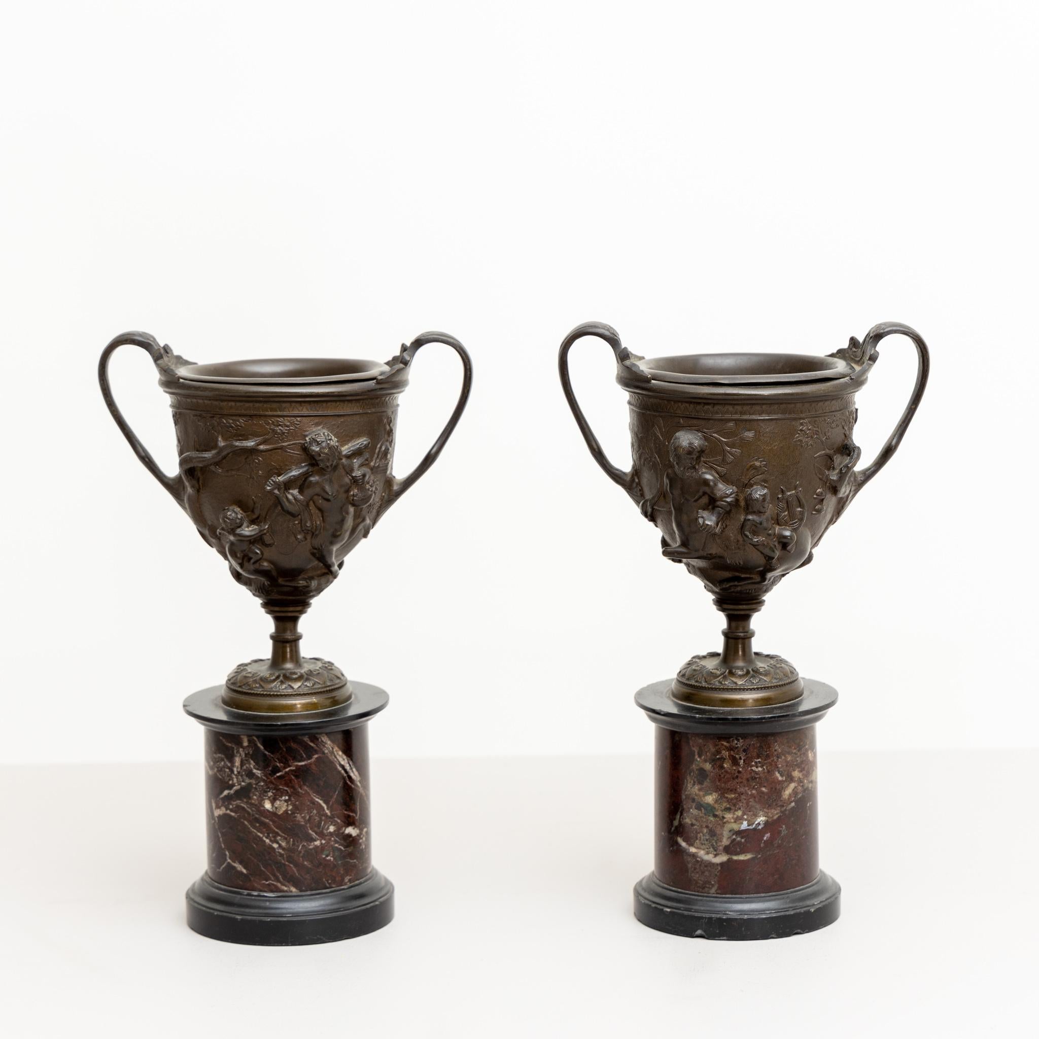 Pair of bronze tazzas on round marble bases with relief depiction of satyrs and heroes on the wall. After a Pompeian original.