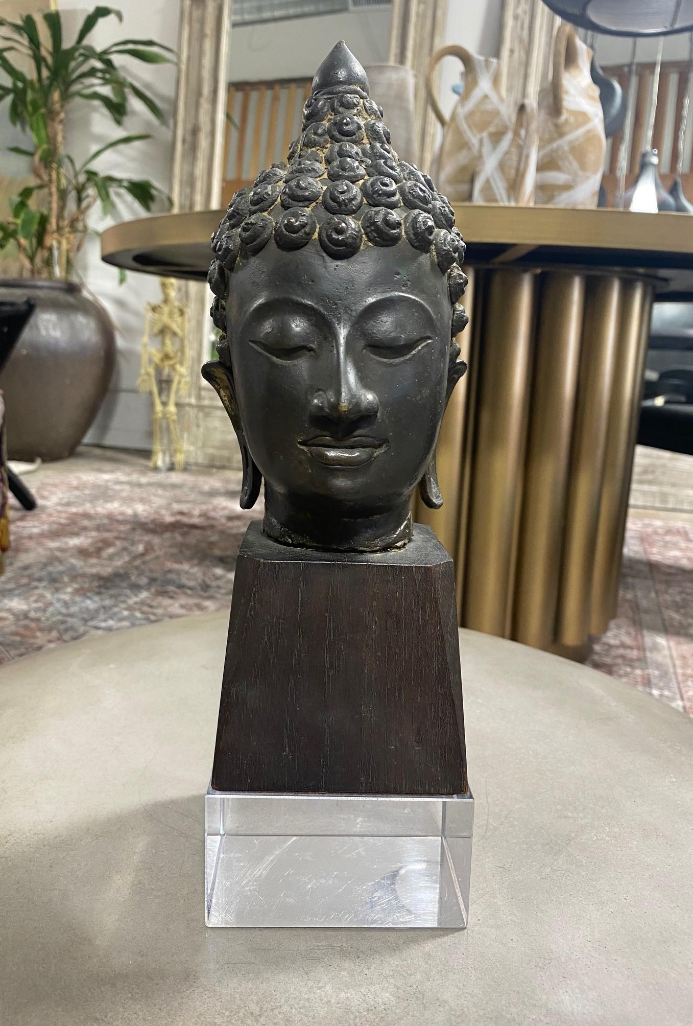 A wonderfully sculpted Thai/ Siam Southeast Asian bronze Buddha head on a custom wood and lucite stand. The Buddha's eyes are closed in serene meditation. The piece has a nice feel and heft to it. Very well well crafted with a gorgeous patina.