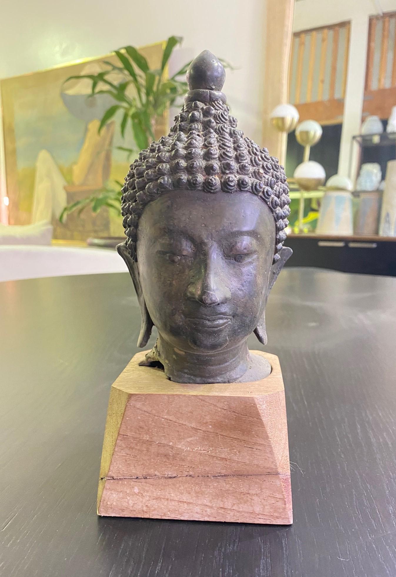 A wonderfully sculpted bronze Thai/ Siam Buddha head on a custom-made wood stand. The Buddha's eyes are closed in serene meditation. The piece has a nice feel and heft to it and fits snuggly into the custom-fitted wood stand. Very well crafted with