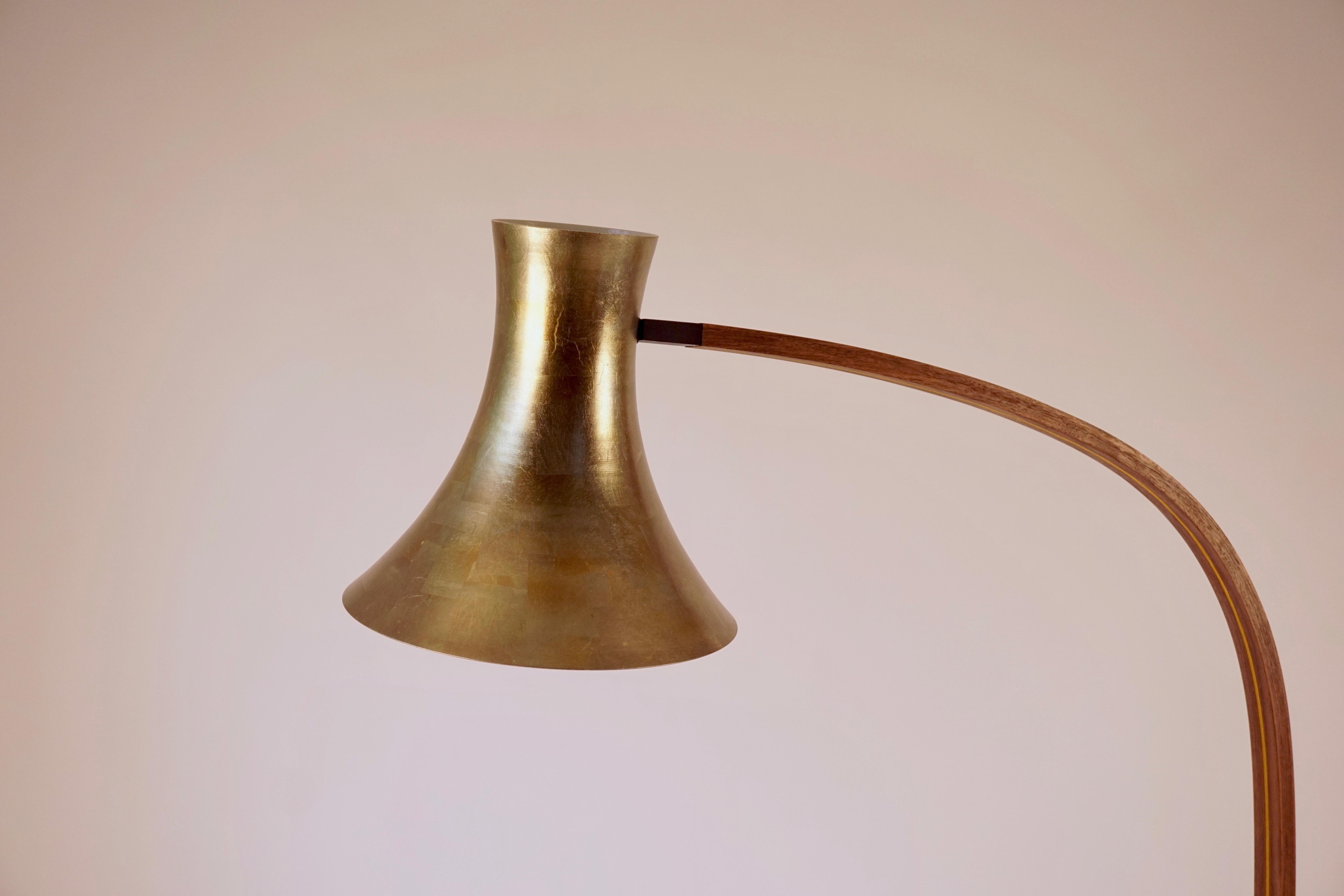 The spun floor lamp features a hand spun aluminum lamp trumpet shaped shade that has been powder coated bone white on the inside. The exterior of the shade has been gilded with a bronze colored leaf. The lamp base and the arm are both walnut, the