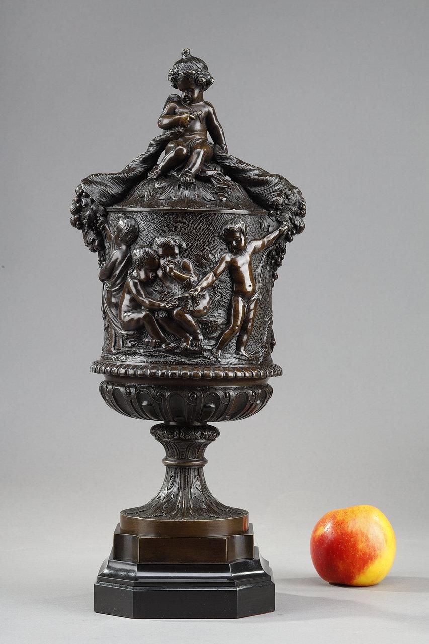 Bedeckte Kanne aus patinierter Bronze im Louis XVI-Stil, im Geschmack von Clodion. Der Hauptteil ist mit einer mythologischen Dekoration aus Putten bedeckt, die durch Weinzweige und Trauben miteinander verbunden sind, die aus mehreren Füllhörnern