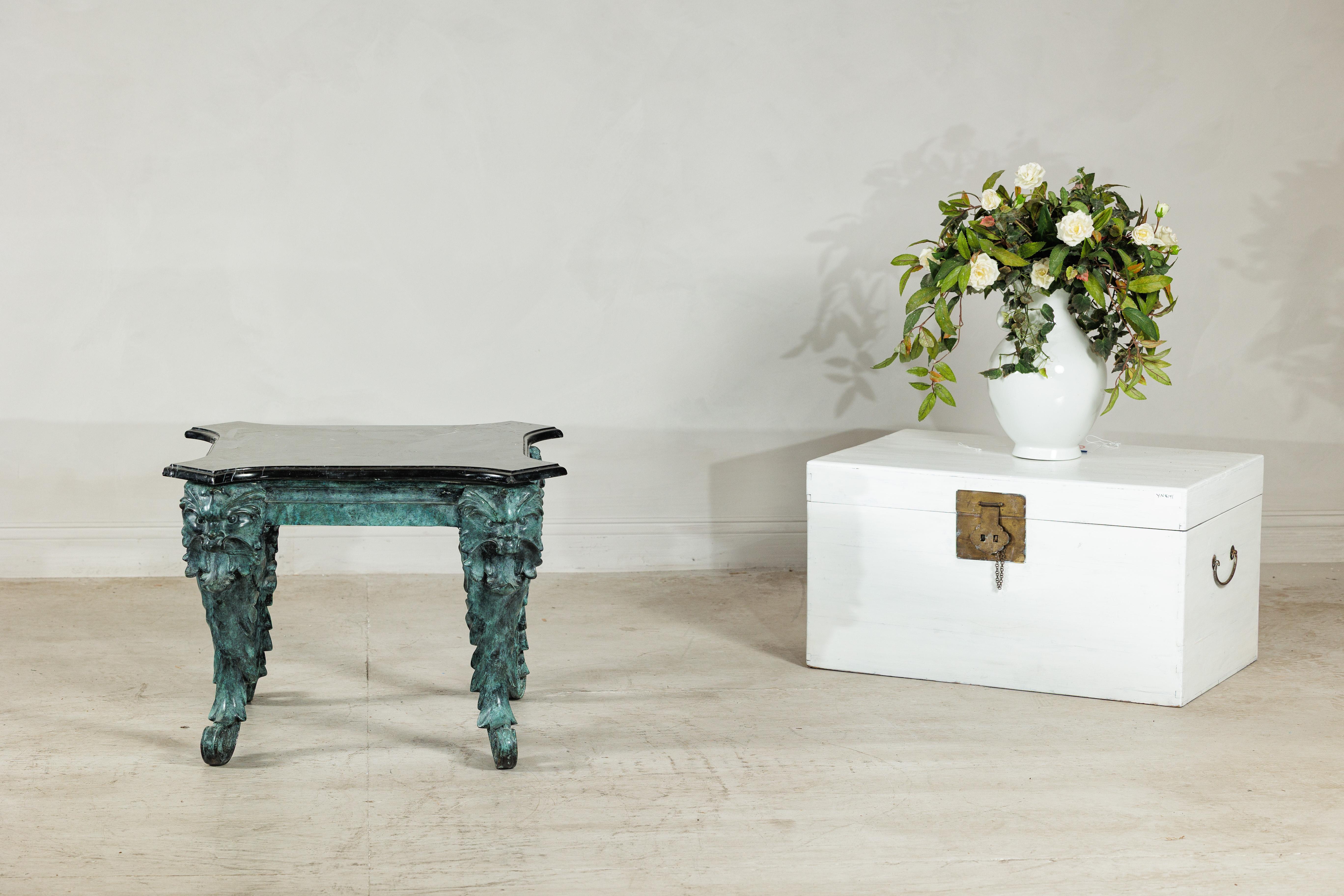 A striking bronze table with a rich verdigris patina and black marble top, its design marked by bold sculptural detail and dramatic presence. The square top features subtly projecting corners and a deep molded edge, resting on four exuberantly