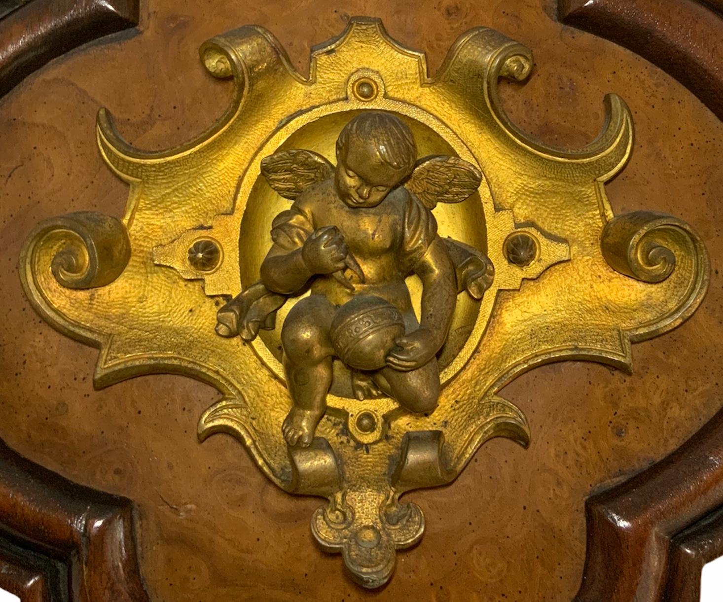 This plaque depicts a bronze winged cherub who is working with a tool in his right hand trying to fix a round object held in his left hand. He is kneeling in one leg over a bronze shaped scrolls border plaque. This in turn stands in a quatrefoil
