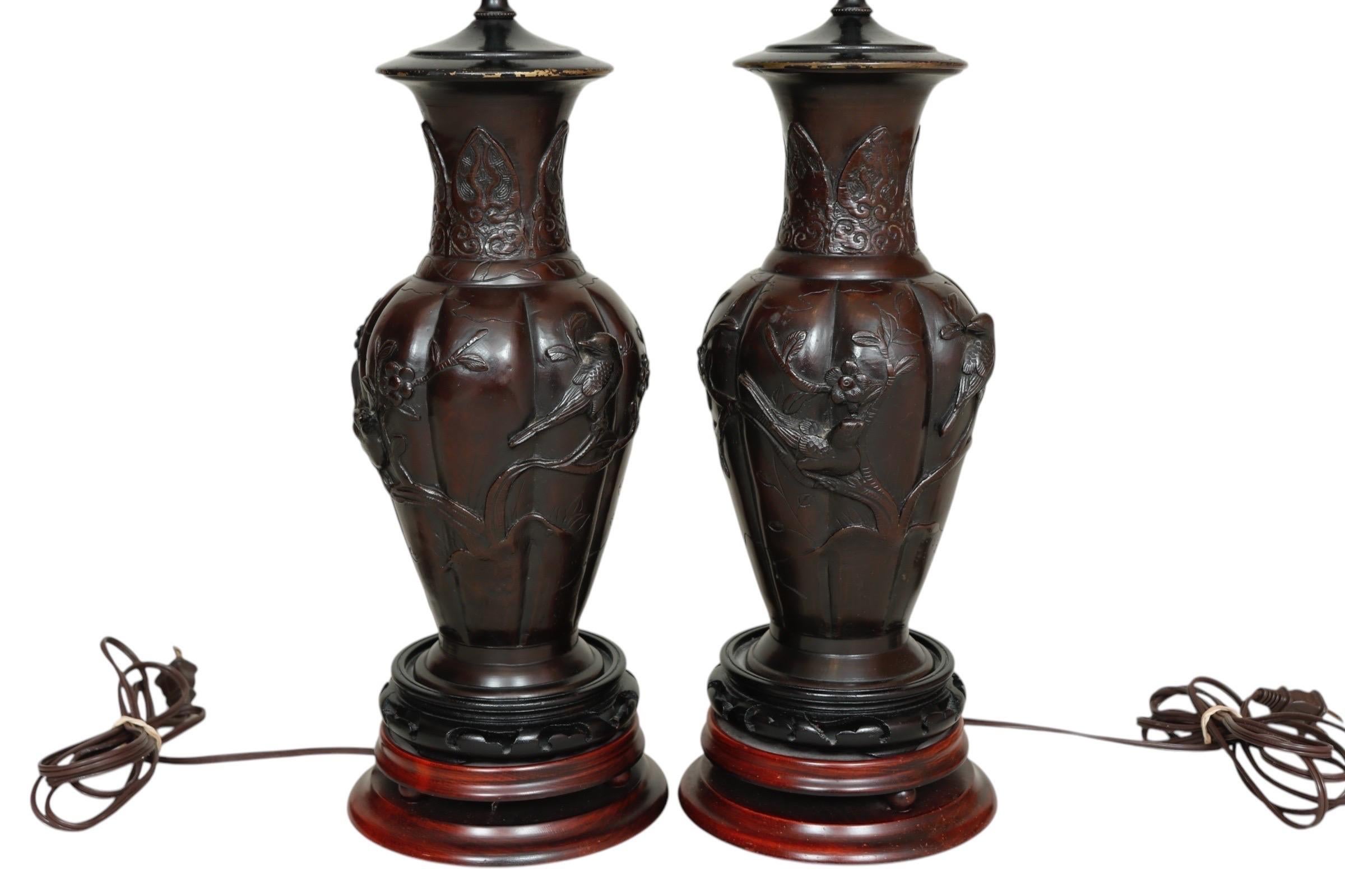 A pair of bronzed Chinoiserie table lamps. Elaborately decorated with small birds in high relief, with cherry blossom branches and small butterflies. The round two tier base is raised up on round feet, beveled and made of rosewood. Each lamp