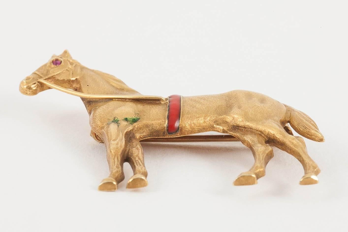 Antique equestrian brooch of a standing racehorse in 14 karat yellow gold set with a ruby eye and a red enamel band around its girth. Single pin fastening.
Measures 32mm in height x 35mm in length.
Antique piece (over 100 years old).
Early 20th