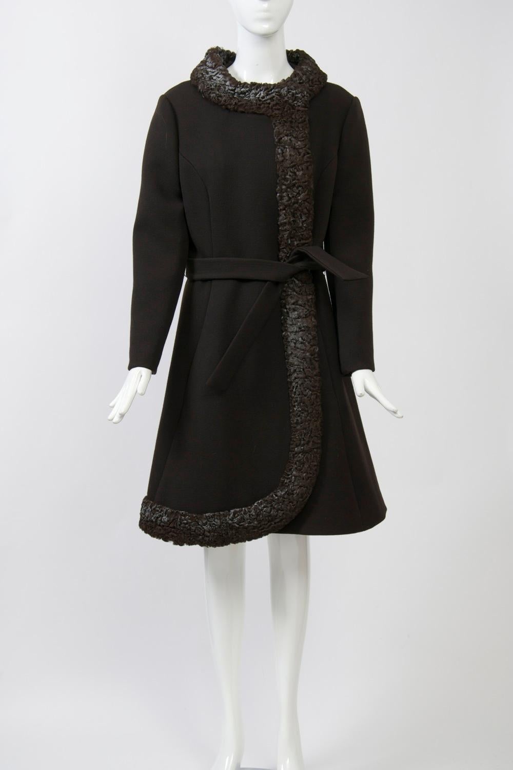 Brown wool coat from the 1960s, its distinguishing feature the mottled brown/gray Persian lamb border that forms its stand-up collar, front edge, and curved knee-length hemline in front. A brown wool tie belt wraps around the seamed waist. Set-in