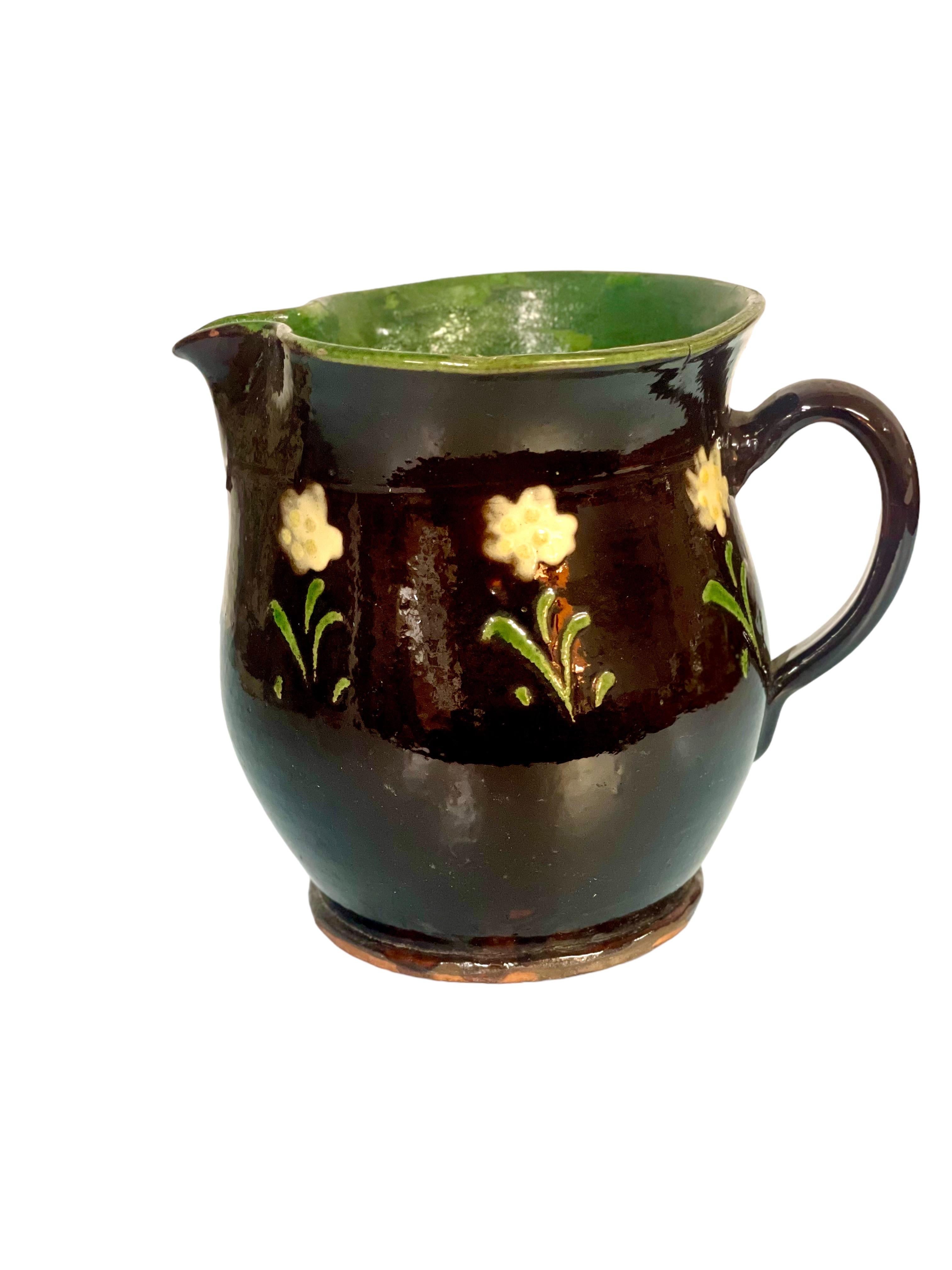 A very attractive little ceramic water or milk jug with handle and shaped spout, its exterior glazed in dark brown and hand-painted with a gorgeous encircling design of flowers, and its interior glazed in vibrant green.