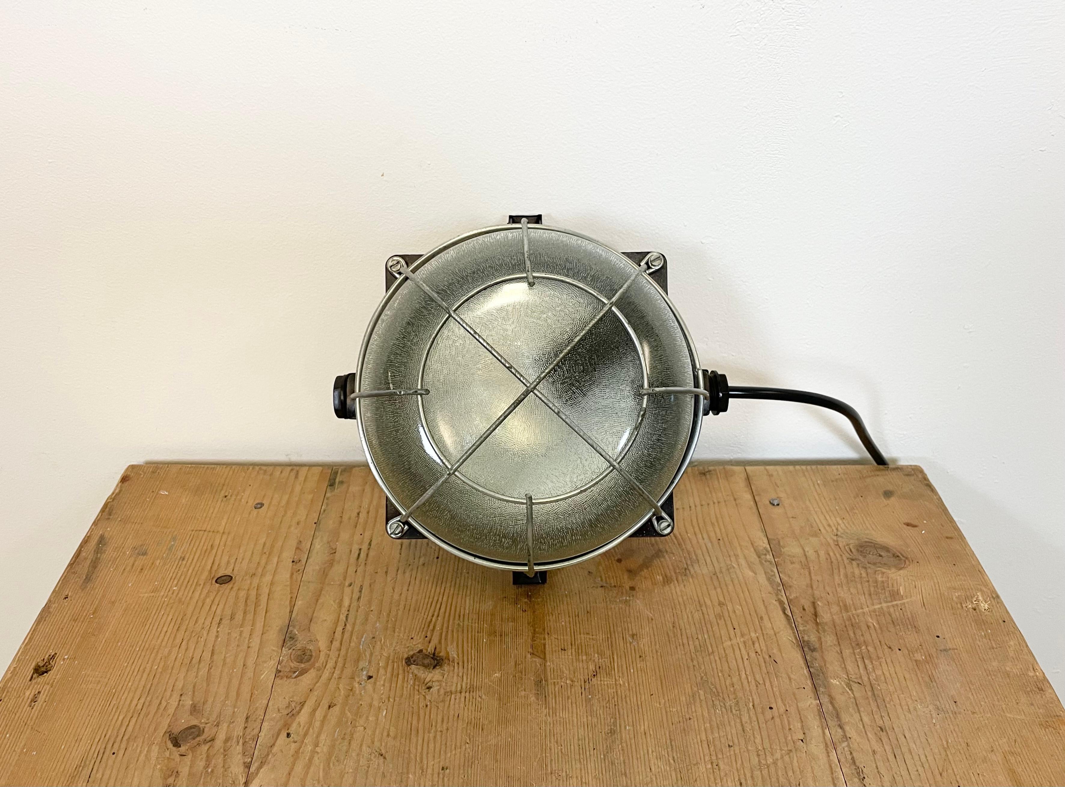 Vintage industrial bakelite wall lamp made in former Czechoslovakia during the 1970s. It features a bakelite body, frosted curved glass cover and iron protective grid. The socket requires E 27 light bulbs. It can be also used as a ceiling lamp.