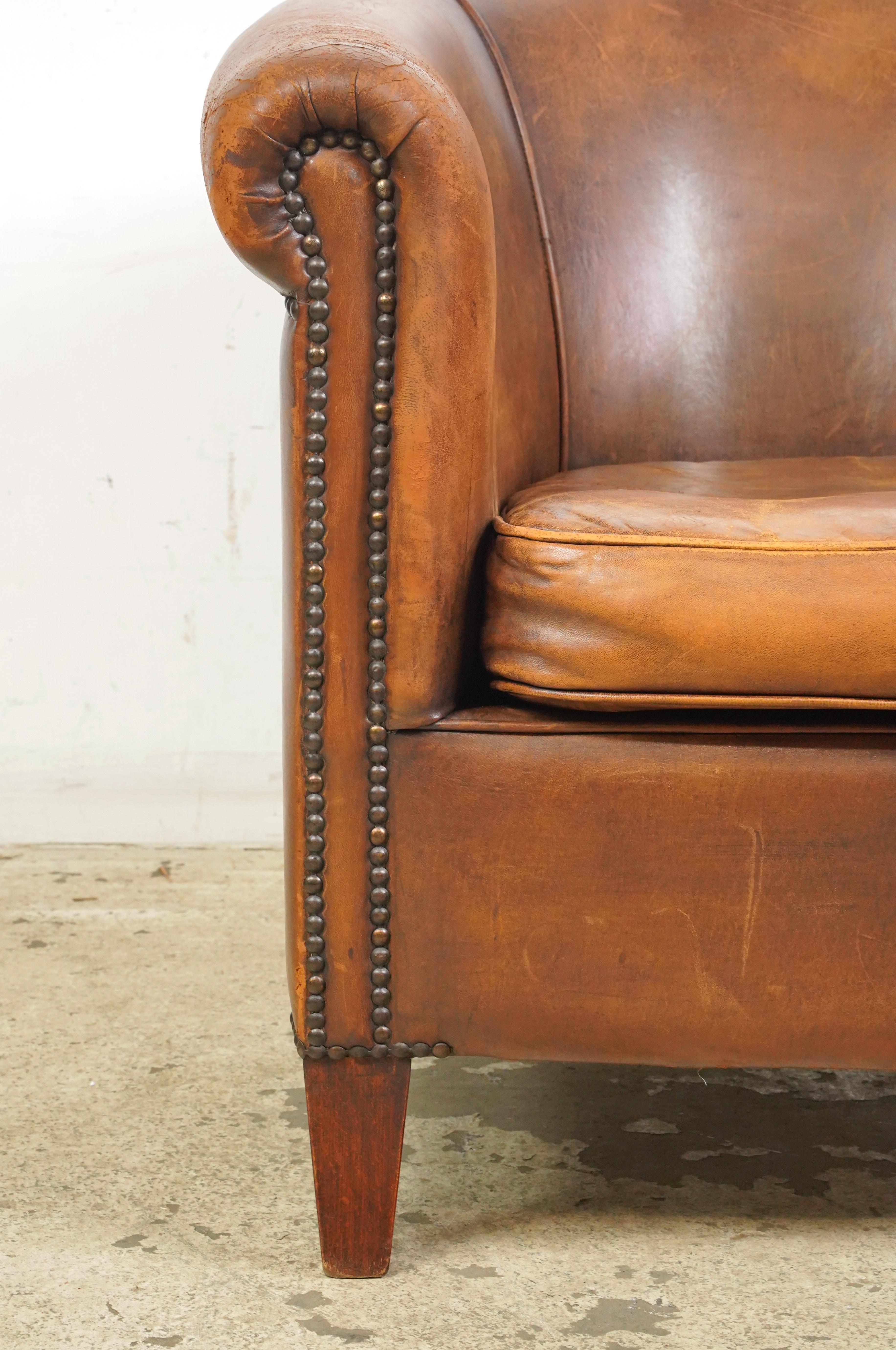 Brown leather studded club chair made in France. It has a round back, wide armrests, a deep cushioned seat, studded details, and wooden legs. The leather and feet show signs of wear and age. Please refer to the pictures for a full view of the