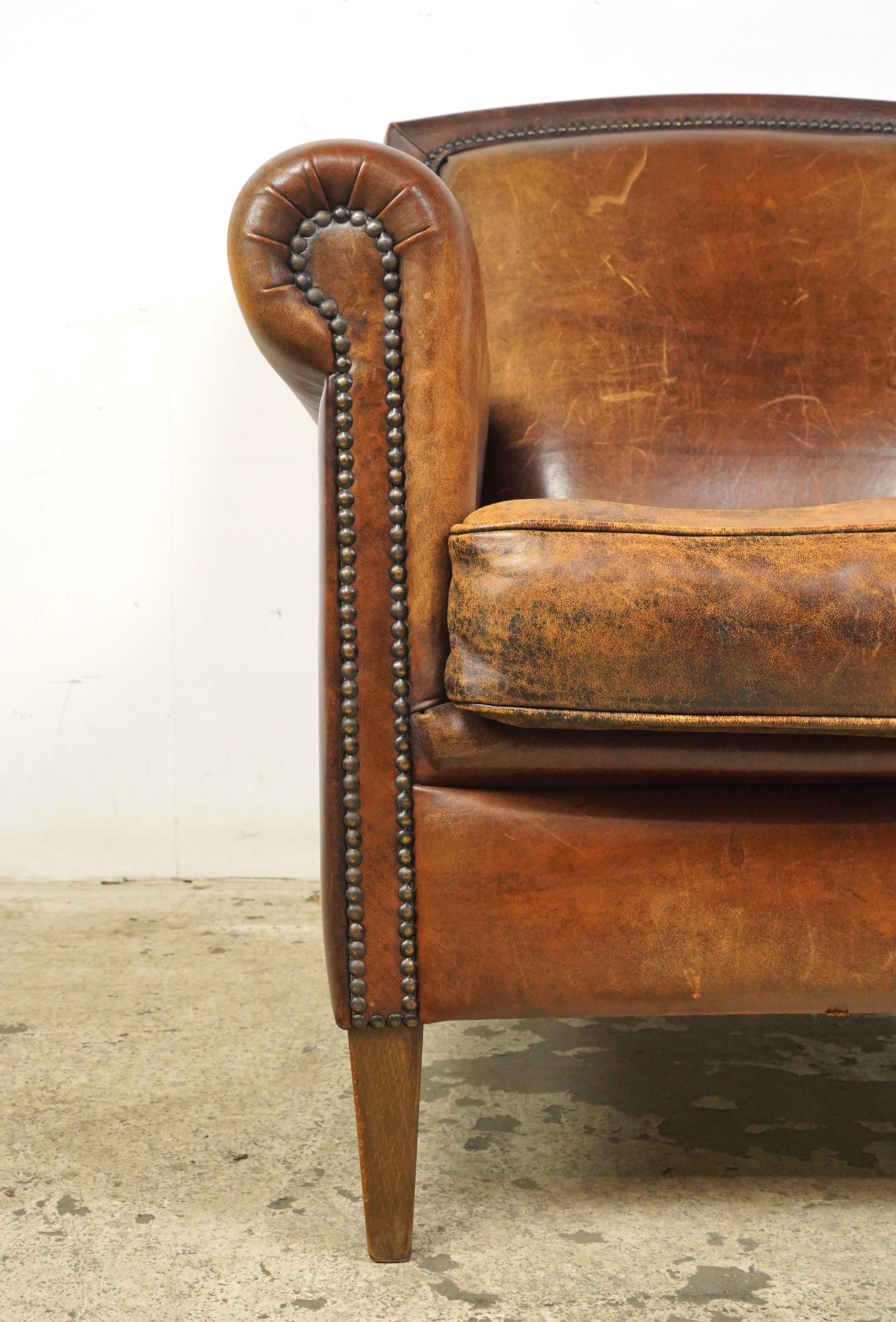 Dark brown leather studded club chair made in France. It has a rounded back and armrests, a cushioned seat, studded details, and wooden tapered legs. The leather and feet show signs of wear and age. Please refer to the pictures for a full view of