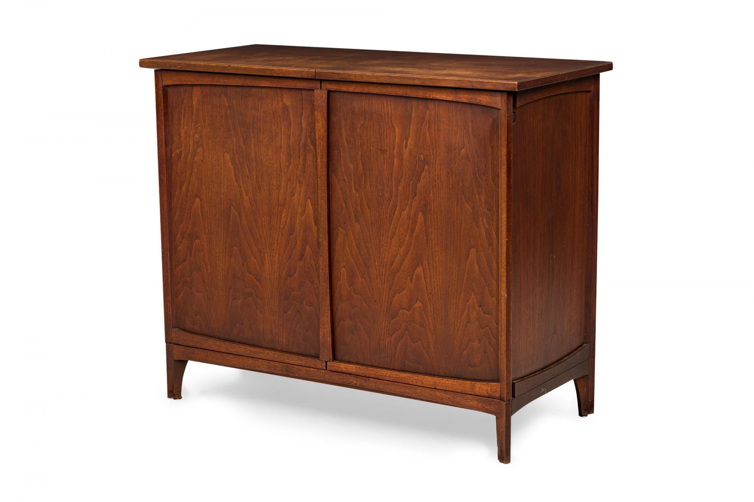 American mid-century walnut dry bar cabinet with a rectangular profile and two doors and a top that all slide outward to reveal an interior compartment with an upper shelf accessible from above, a central drawer, and an open lower cabinet, resting