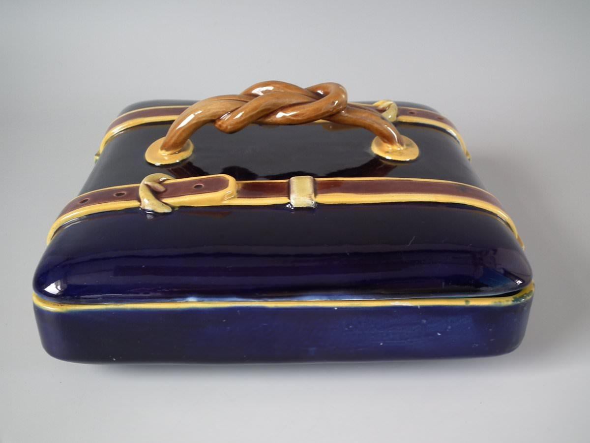 Brown Westhead Moore Co. Majolica box and cover which features a suitcase (perhaps), with two belt straps either side of a twisted rope handle. Coloration: cobalt blue, brown, ochre, are predominant. Bears a pattern number, '609'. Book reference,