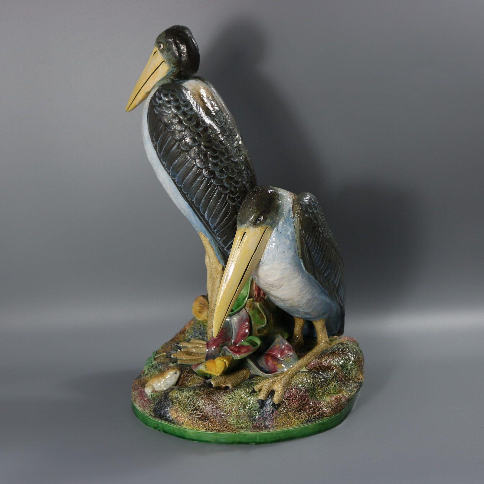 Brown Westhead Moore and Co Majolica figural vase which features two storks, one standing and the other crouched beside. A frog hides on the ground below. There are leaves and foliage around the base and a tree stump forms the vase at the back.