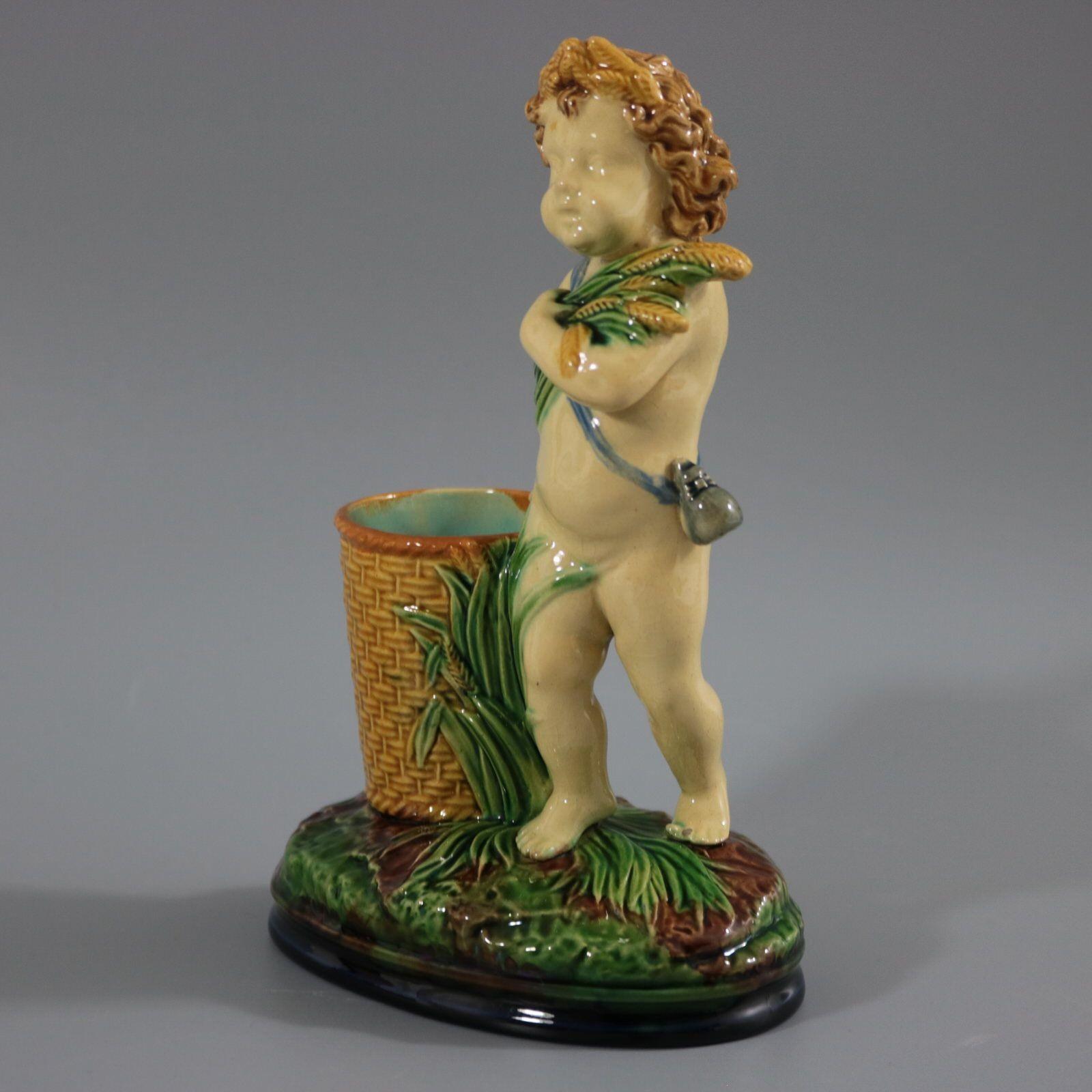 Brownfield Majolica posy holder which features a putti carrying a bunch of wheat. Stood beside a wicker basket. Colouration: green, cream, yellow, are predominant. The piece bears maker's marks for the Brownfield pottery. Bears a pattern number,
