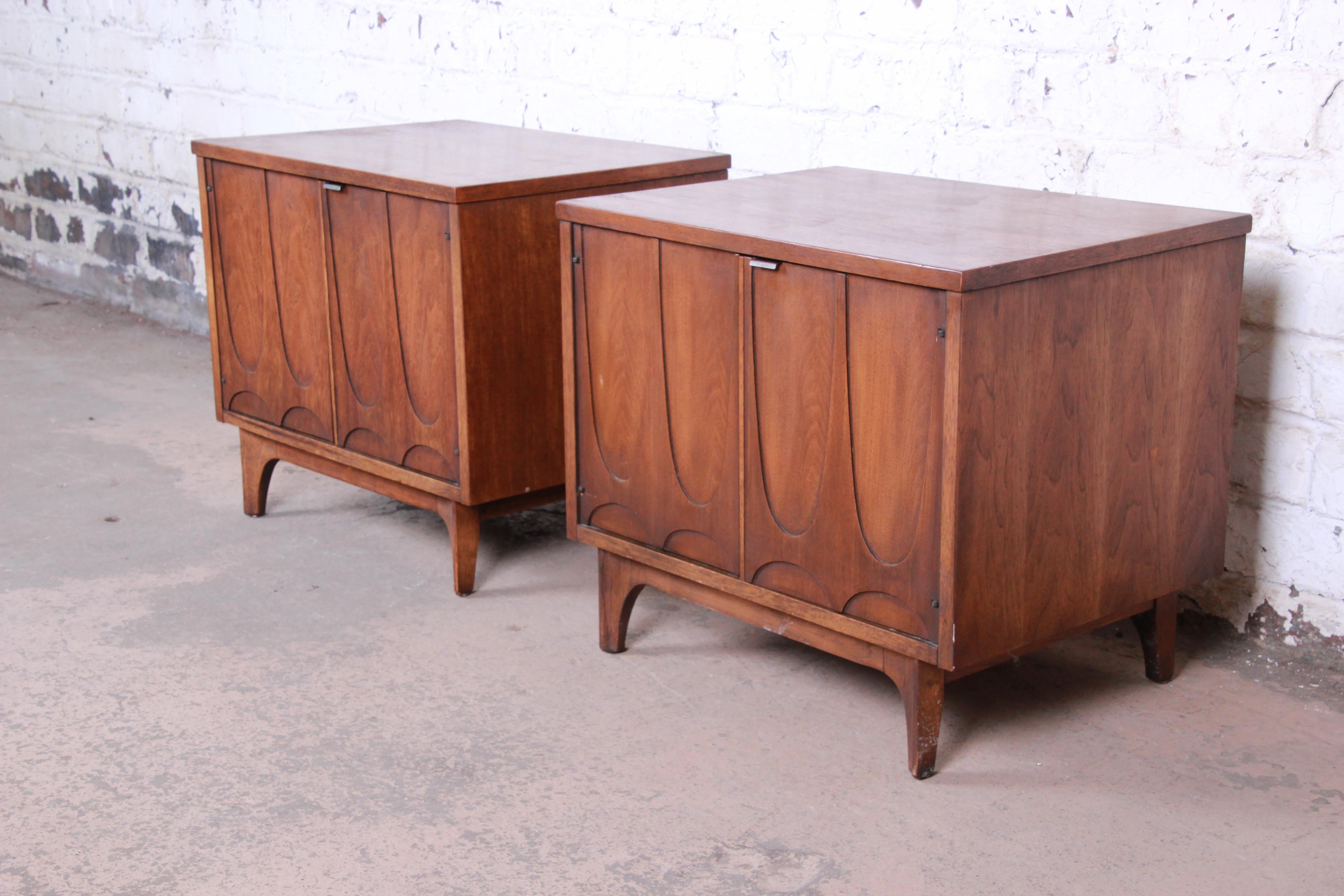 A stunning pair of Mid-Century Modern sculpted walnut nightstands or side tables by Broyhill Brasilia. The nightstands feature beautiful walnut wood grain and an iconic sculpted arch design. They offer good storage, each with an open cabinet space
