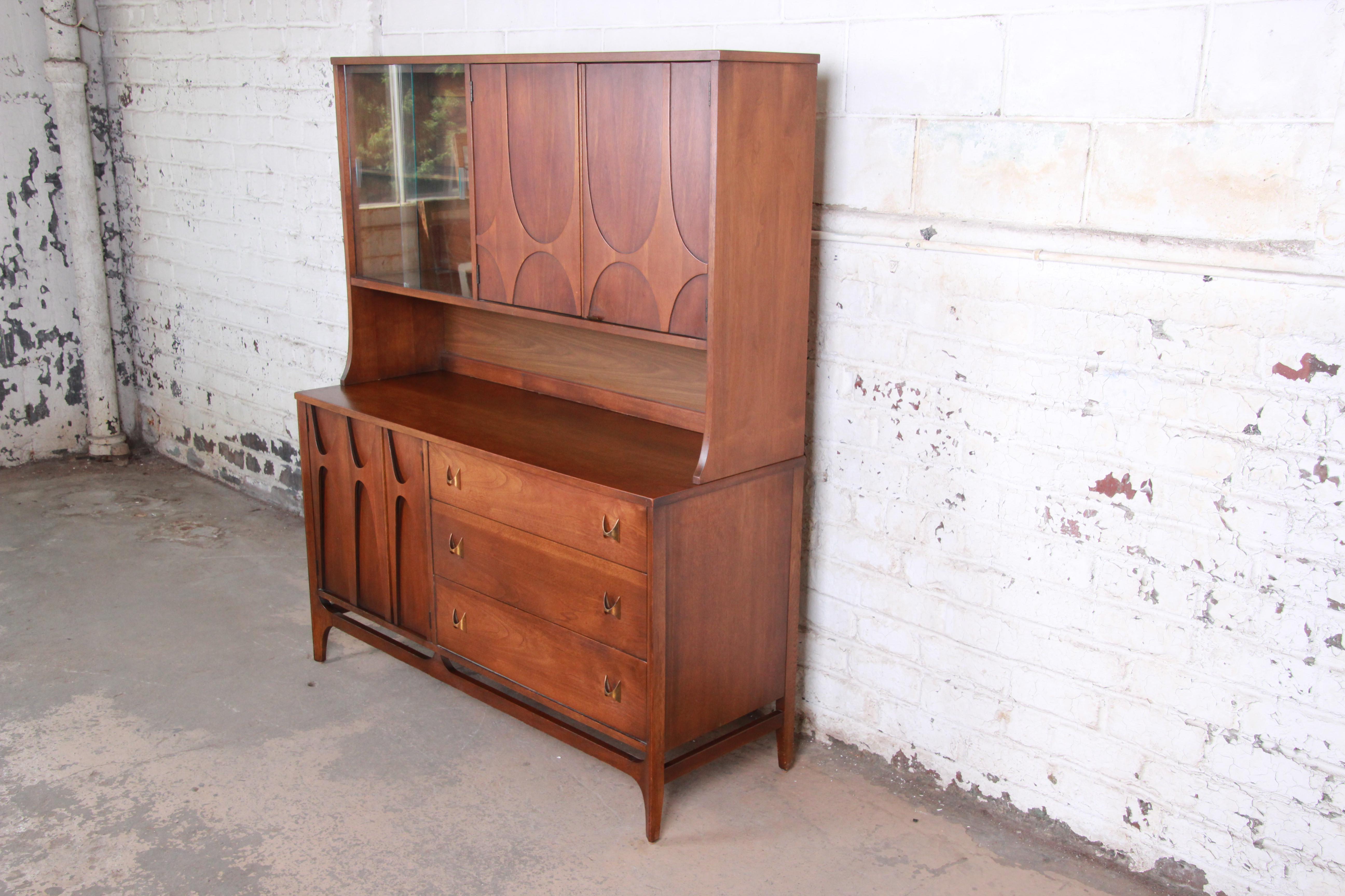 An exceptional Broyhill Brasilia mid-century modern sculpted walnut sideboard with hutch. The sideboard features gorgeous walnut wood grain, with sculpted arches and original brass pulls. It offers ample room for storage with three deep dovetailed