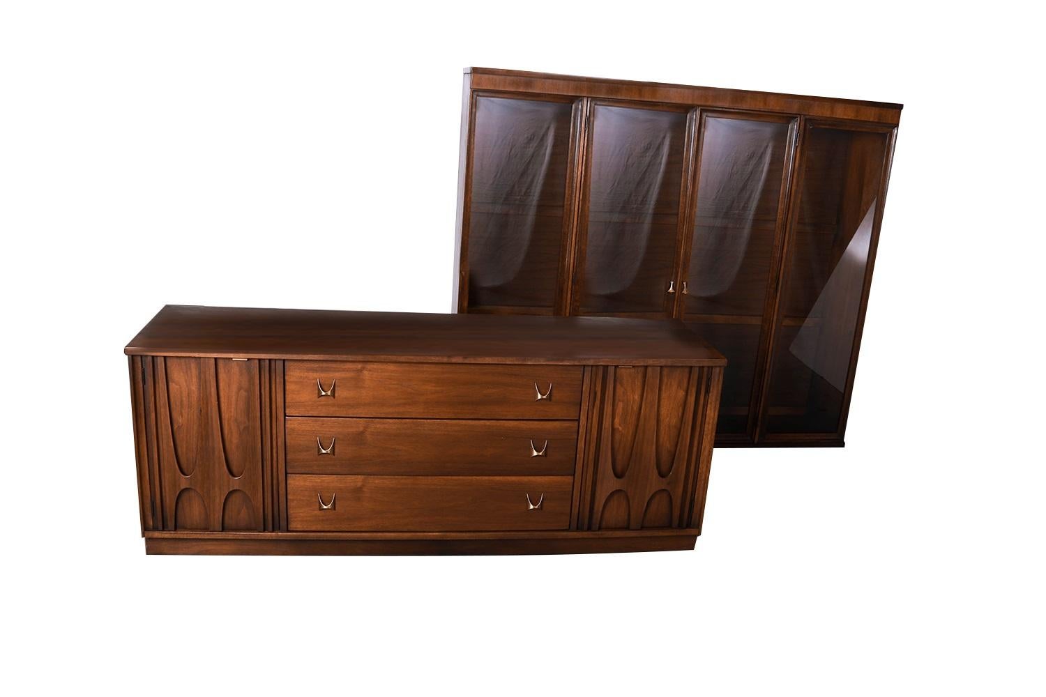 A beautiful Broyhill Brasilia Mid-Century Modern sculpted walnut credenza/sideboard with detachable lighted hutch. Hutch top lifts off the lower cabinet. Upper portion features a lighted cabinet with convenient glass doors that open to reveal two