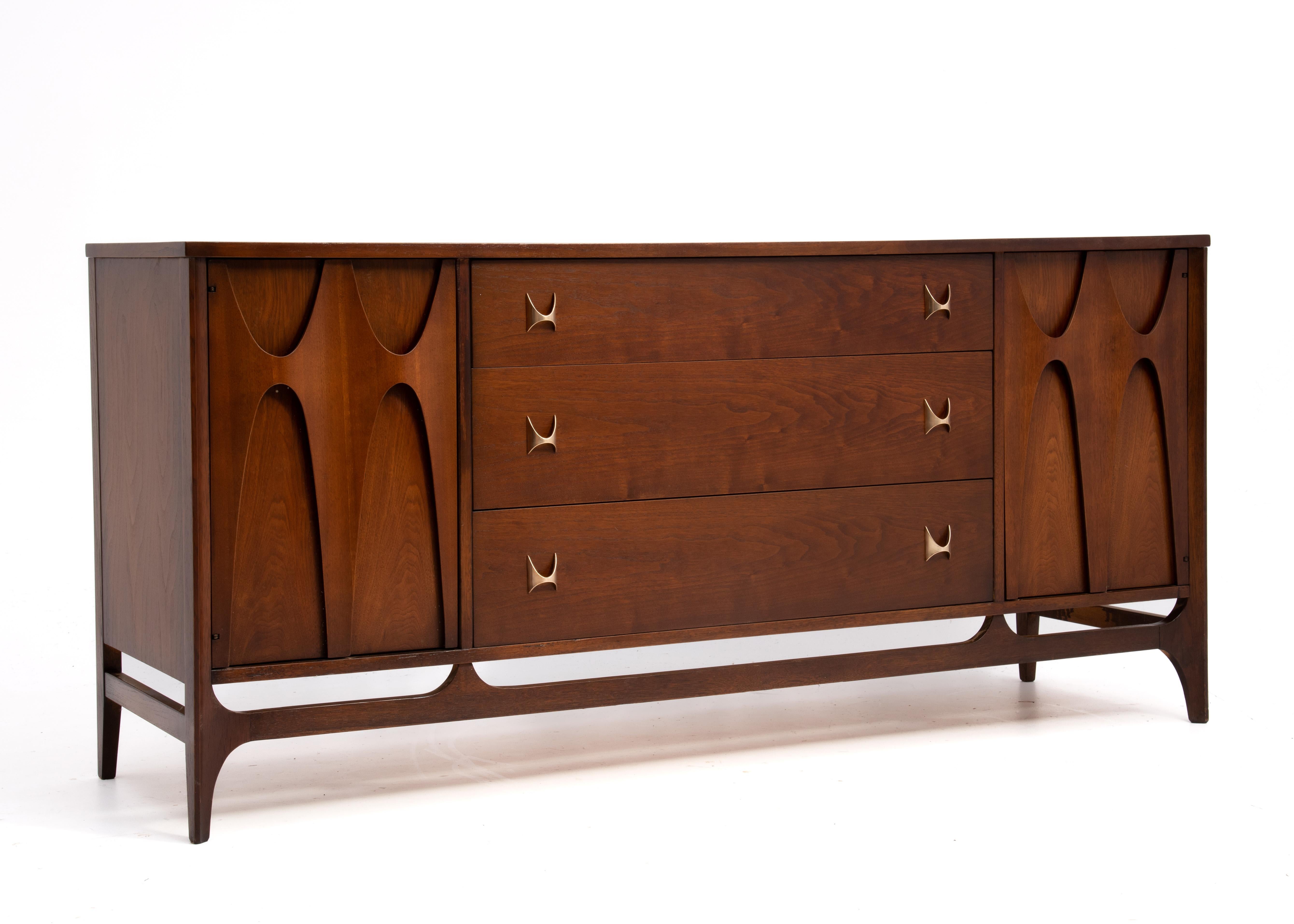 A Mid Century Modern Broyhill Brasilia three drawer walnut credenza. Two side cabinet doors and brass hardware. Its iconic design includes bentwood swoops on the doors and brass drawer pulls that were inspired by the mid-century architecture of the