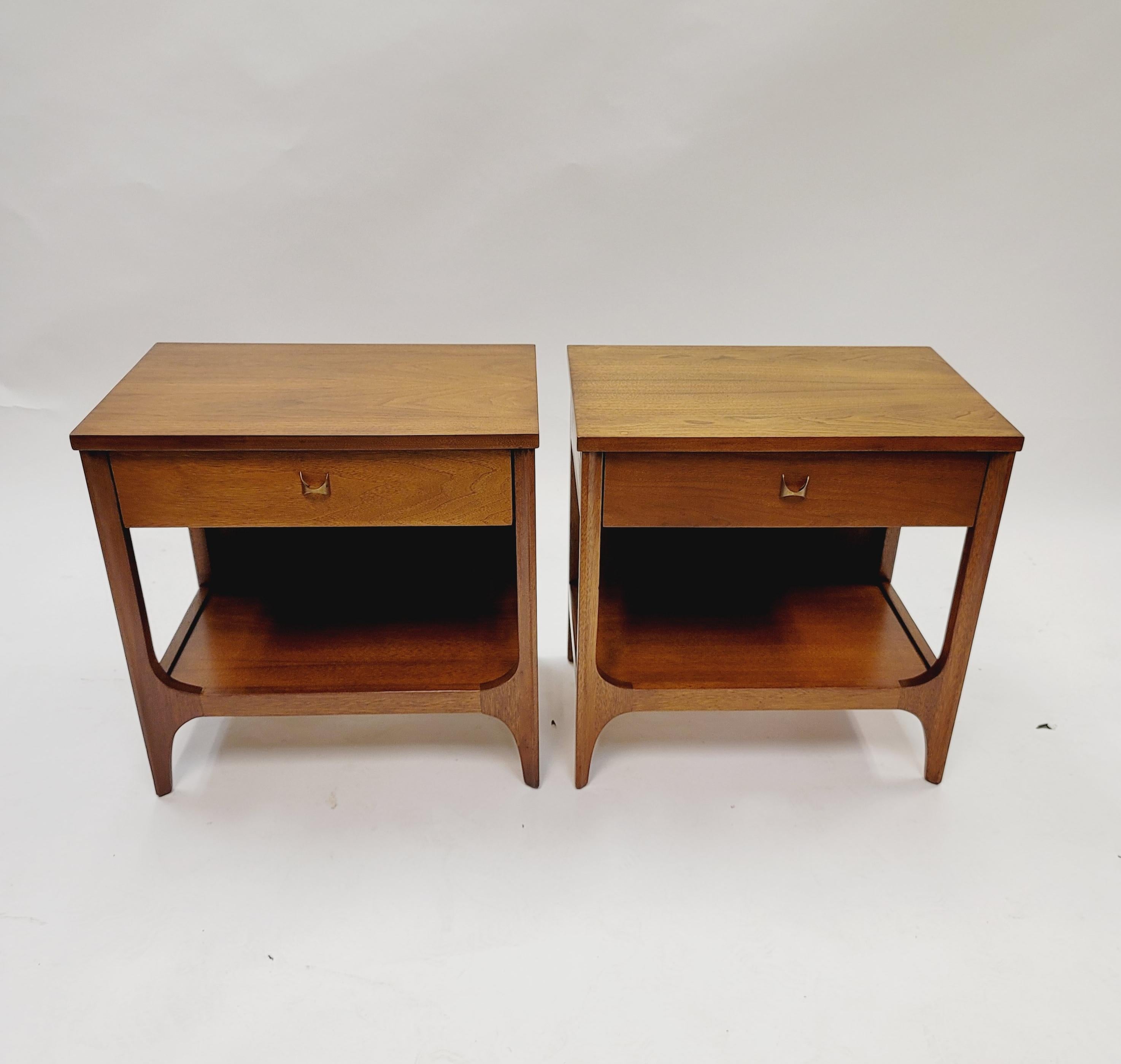 This pair of hard-to-find Broyhill Brasilia nightstands are beautifully constructed from walnut with brass sculptural pulls. One drawer and a lower shelf on each stand provide maximum storage.