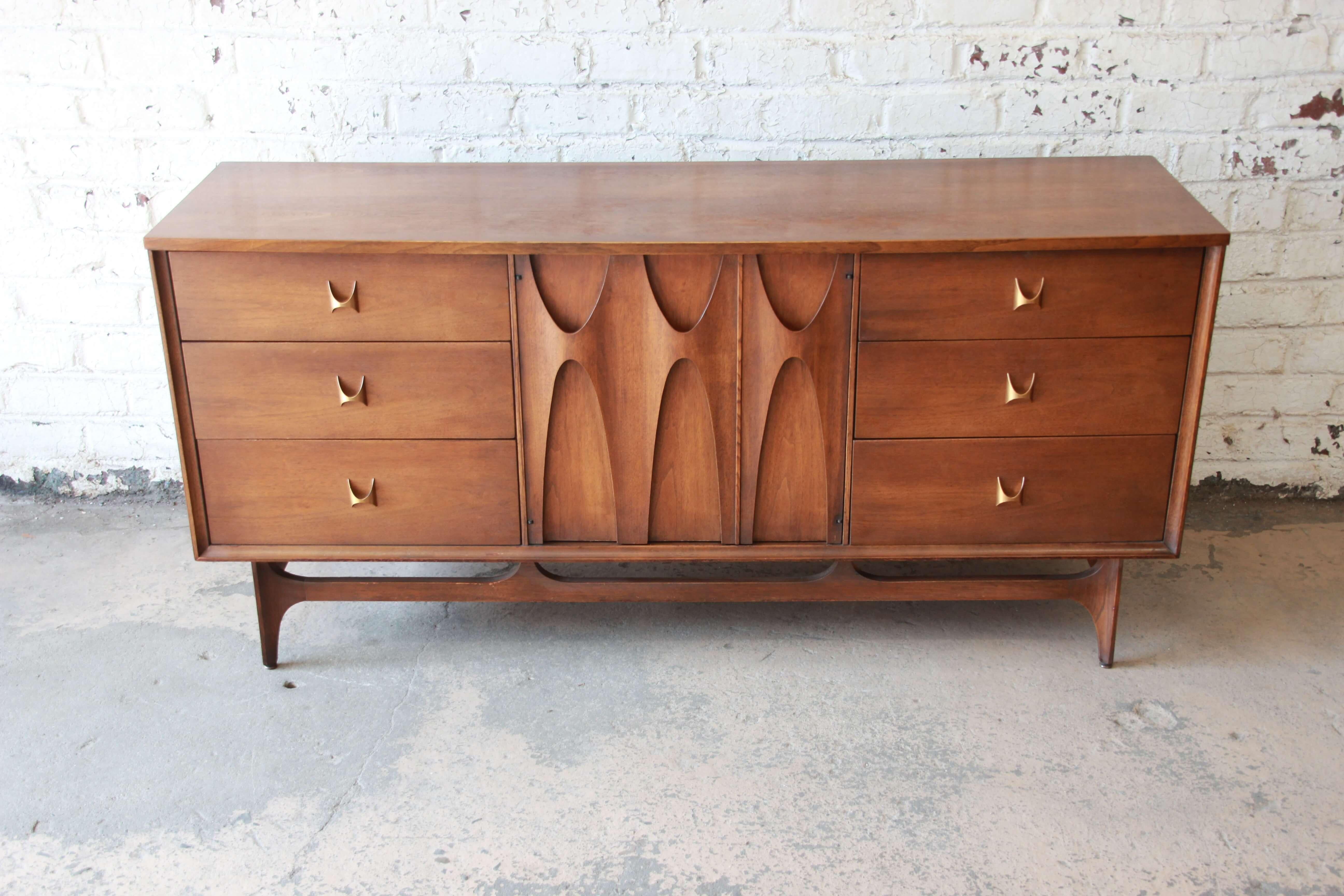 Offering an awesome Broyhill Brasilia sculpted walnut nine-drawer dresser or credenza. This piece has unique brass pulls on each drawer with the three smooth sliding drawers on each side. The center has sculpted arched pulls that are on two cabinet