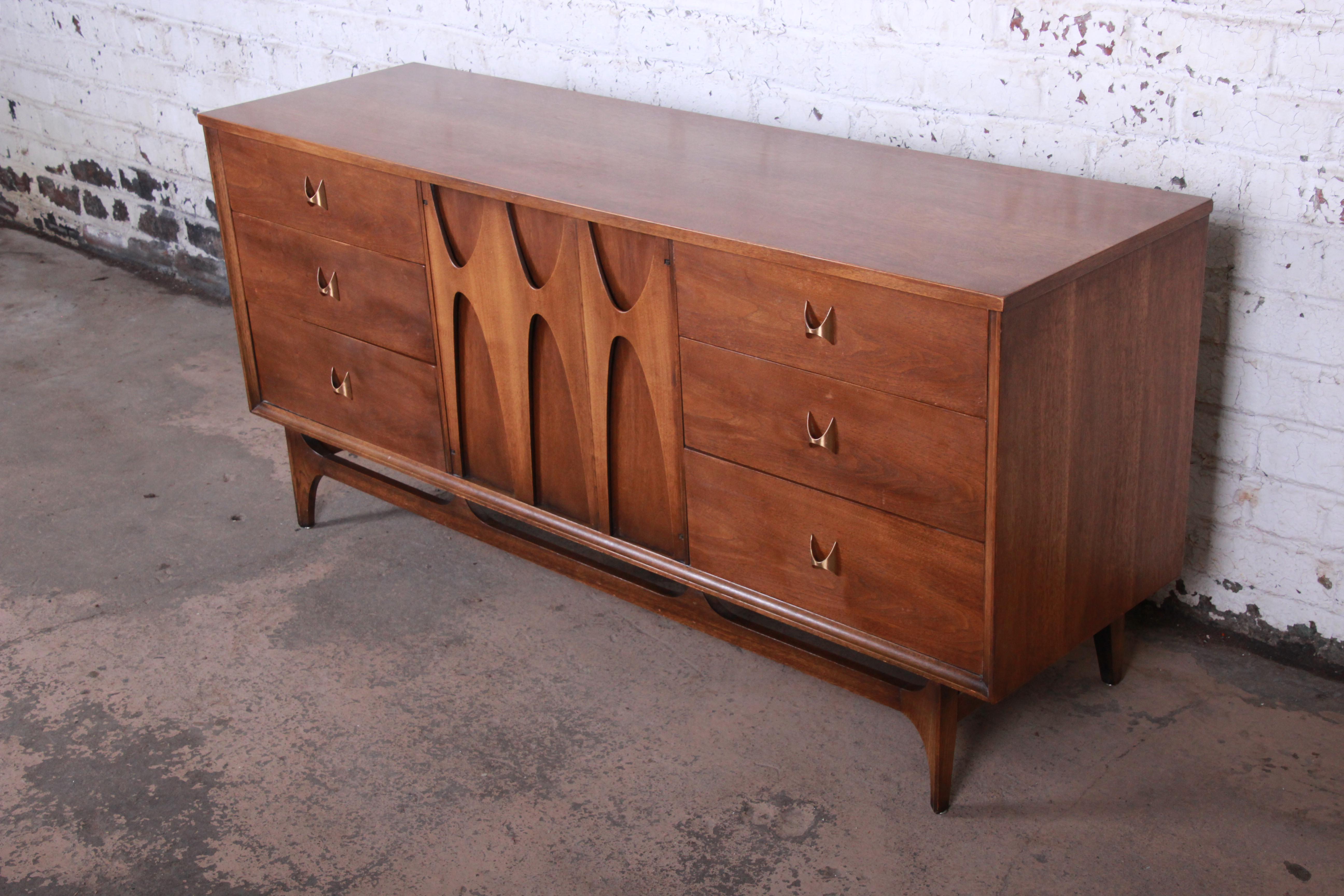 A gorgeous Mid-Century Modern sculpted walnut triple dresser or credenza by Broyhill Brasilia. The dresser features beautiful walnut wood grain, with a unique sculpted design and iconic sculpted arch brass drawer pulls. It offers ample storage, with