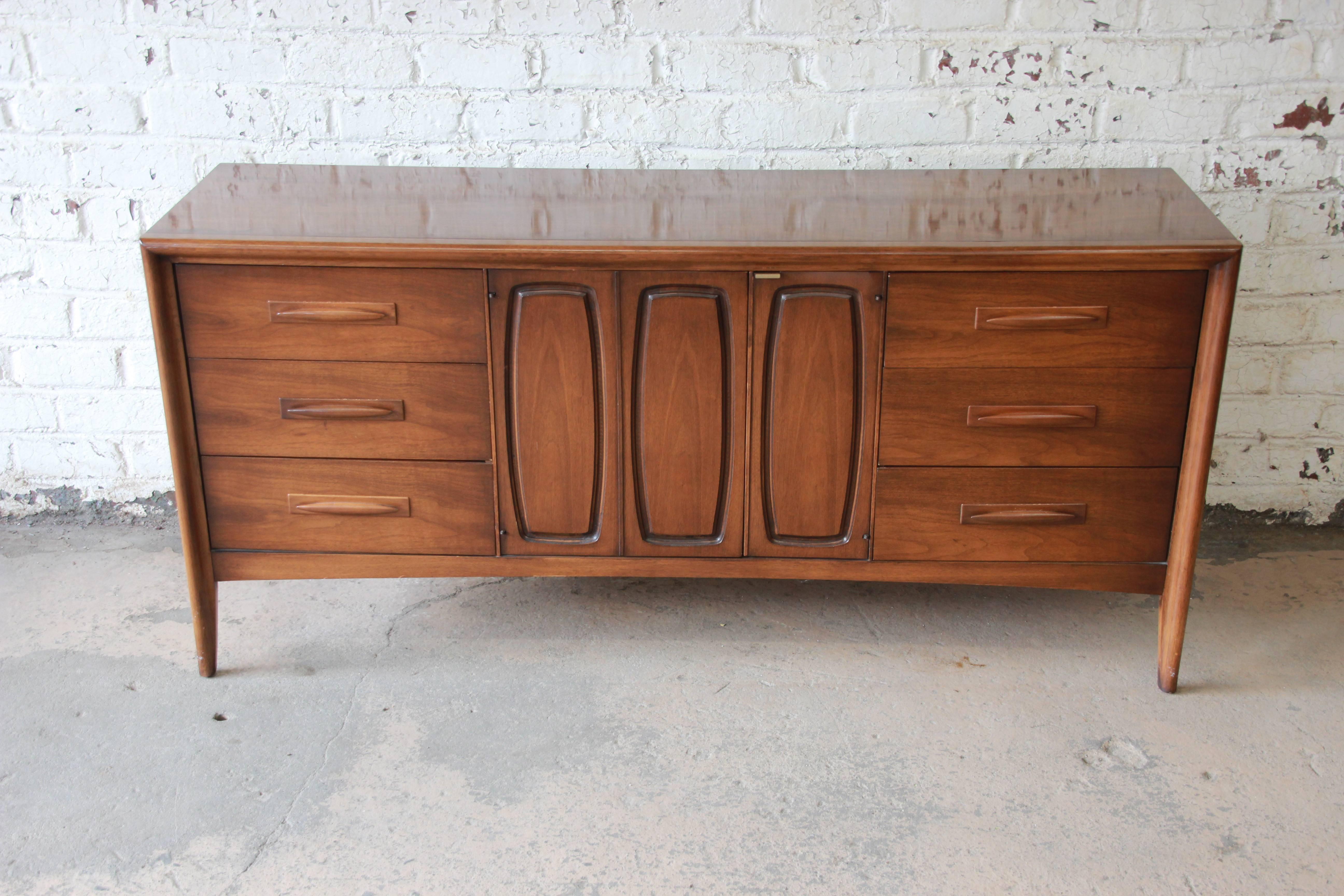 Offering a very nice Broyhill Emphasis Mid-Century Modern sculpted walnut credenza or triple dresser. The credenza has clean midcentury lines, beautiful walnut wood grain, and nice sculpted walnut drawer pulls. It offers ample room for storage, with