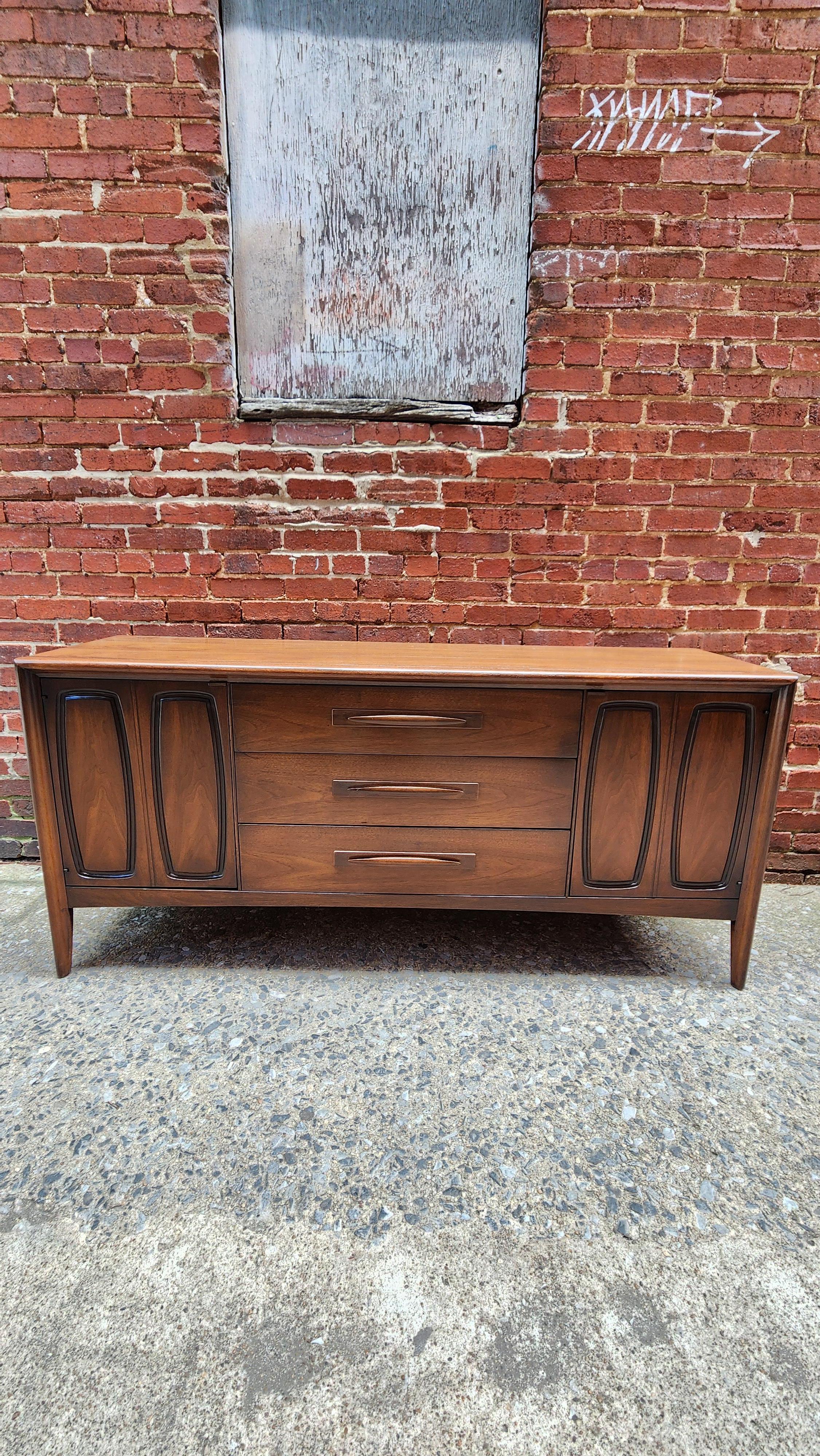 This mid-century modern walnut credenza is from the popular Emphasis line by Broyhill. Crafted at the height of the modernism design eta of the 50s, the Emphasis line combined graceful legs with sculpted walnut pulls and raised panel doors. 
This