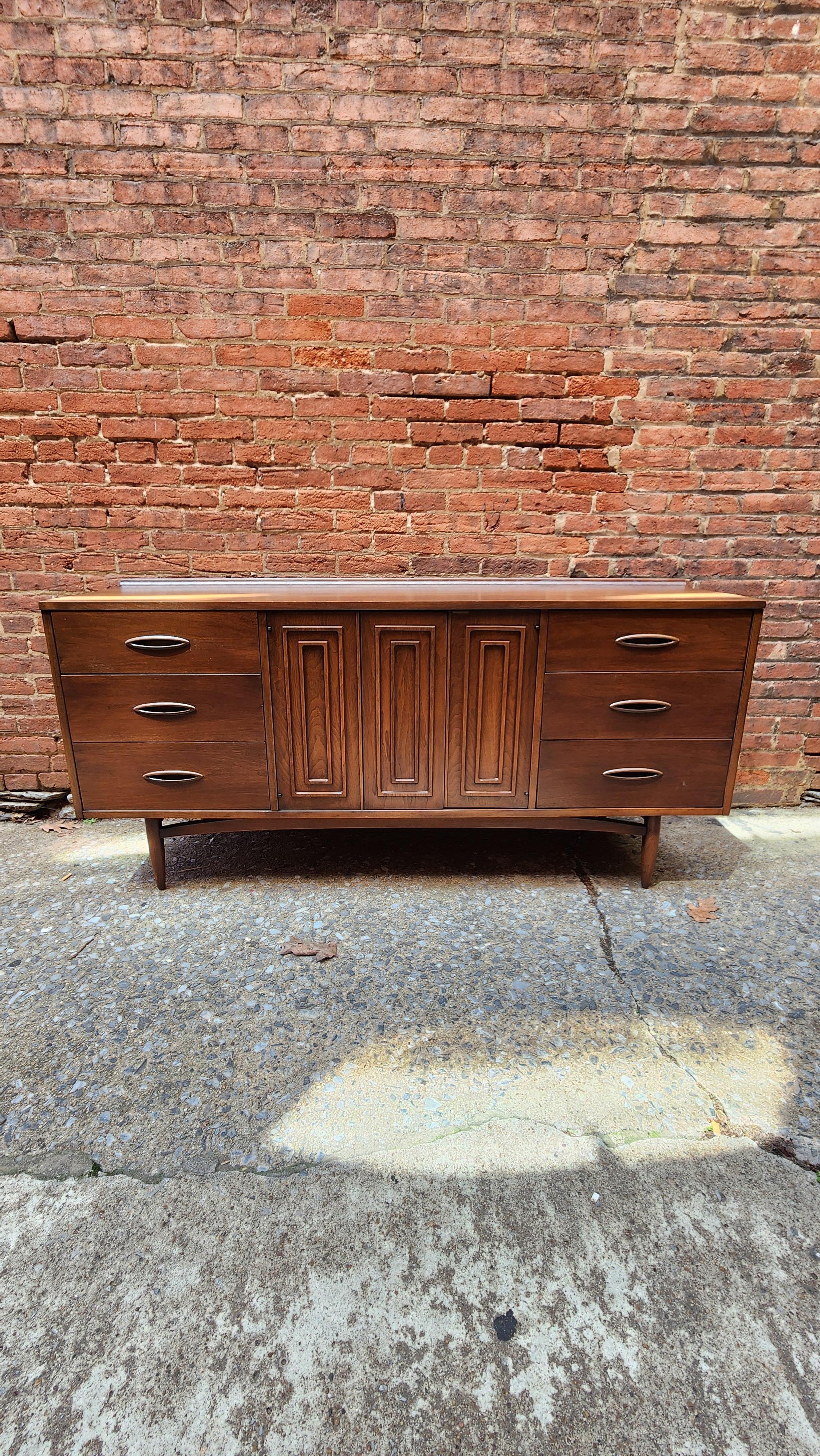 This lovely walnut Mid-Century Modern nine-drawer dresser is from the Sculptra line by Broyhill. This dresser has an additional set of drawers behind each set of folding doors. 
The top has a delicate curve at the back giving it an extra deviously