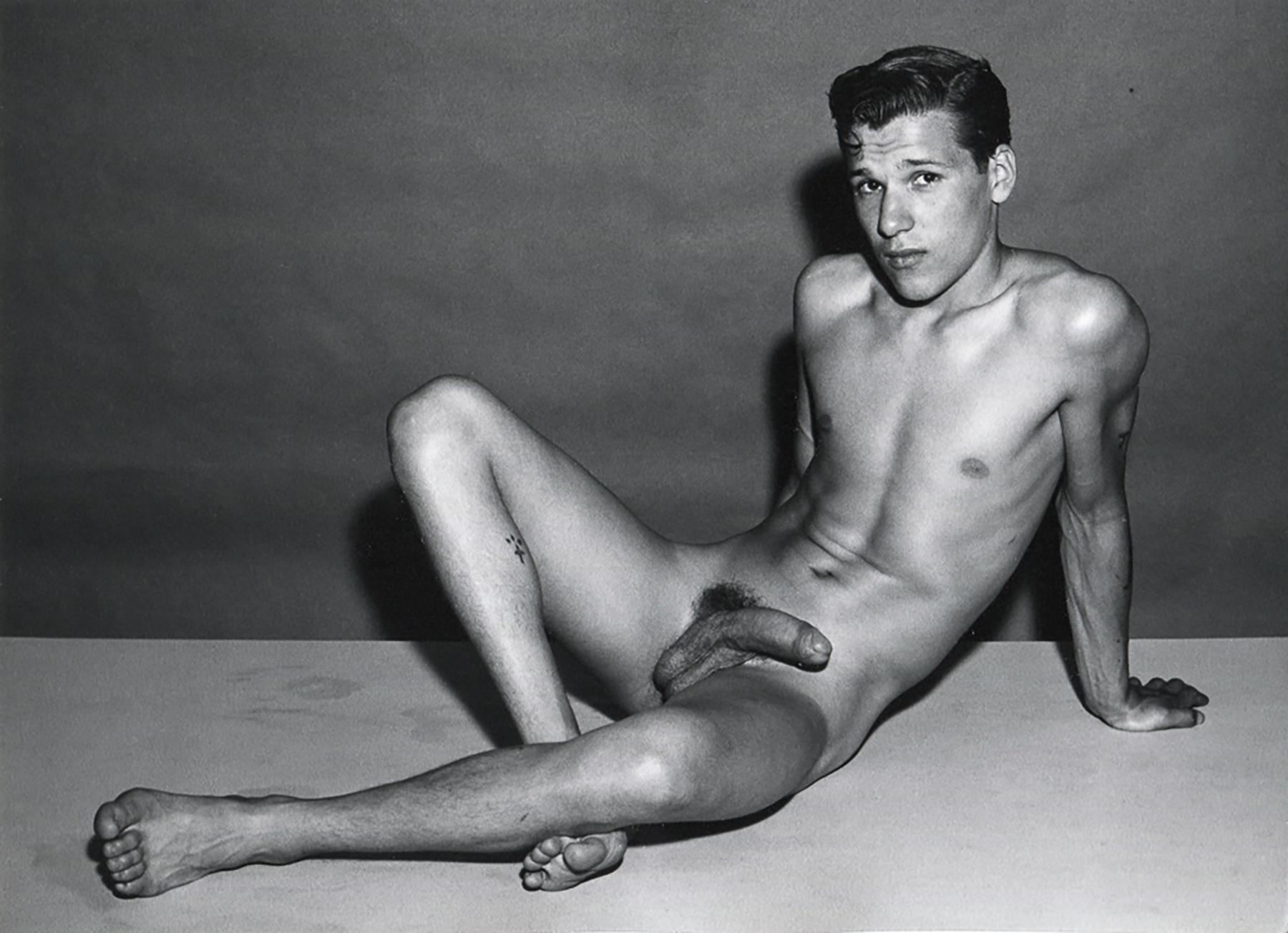 Black and white photograph by Bruce of Los Angeles of a nude male model, Jim Manning, reclining on a flat surface, gazing towards the camera with a confident yet relaxed expression. 

Jim Manning (Reclining Arms Behind Back)
Printed after