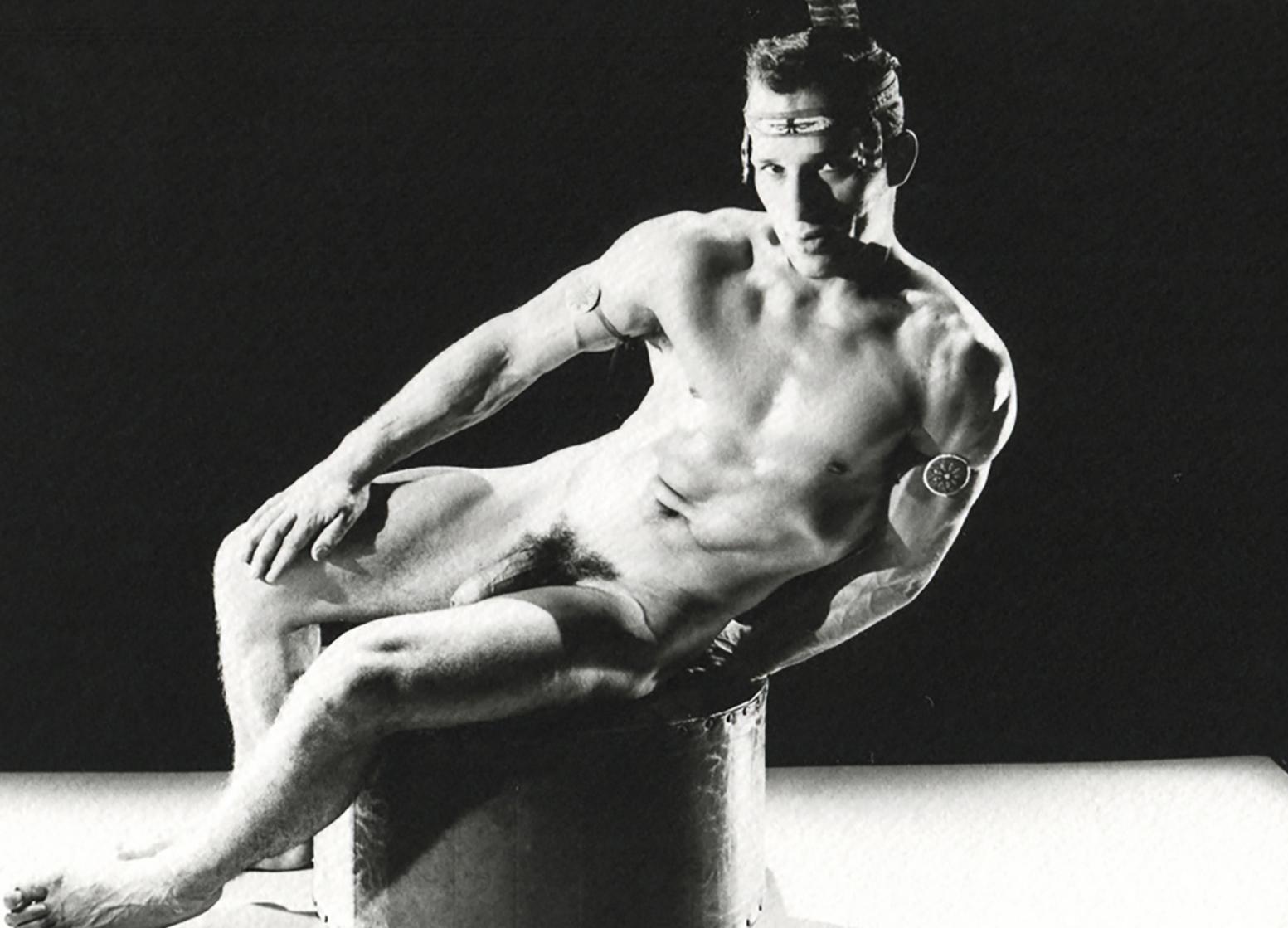 Black-and-white photo by Bruce of Los Angeles (Bruce Bellas) of a nude male figure in a dramatic pose with headpiece and armband, highlighted by stark lighting that emphasizes his muscular form.

Untitled #3245-5 (Man Reclining with Feather
