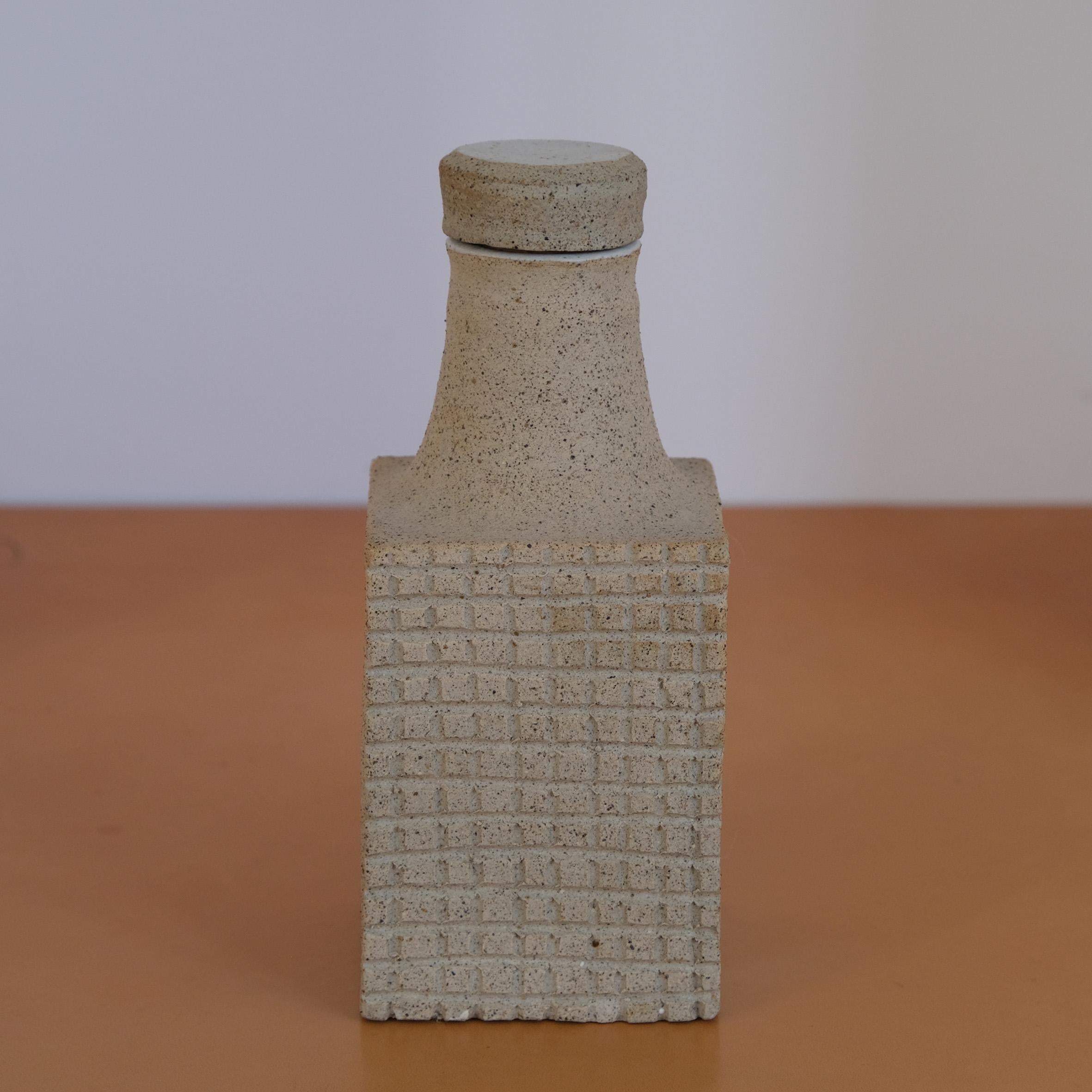 Bruno Gambone (1936-2021)

Flasche

Eine hellgraue Keramikflasche mit einem rechteckigen Körper, der auf der Vorder- und Rückseite mit einem Flachreliefgitter verziert ist. Der trompetenförmige Flaschenhals ist mit einer passenden Keramikkappe