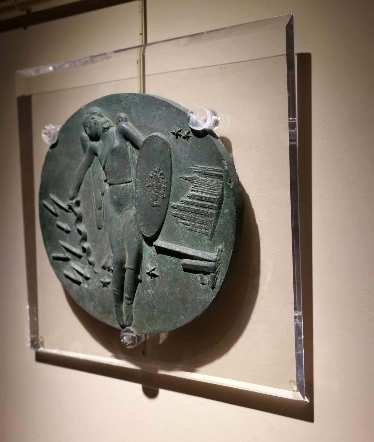 This bronze relief ( 29 x 29 cm, plexiglass pedestal 41 x 41 cm) is a model for a commemorative medal dedicated to the Italian Air Force. 
At the centre there is the Allegory of the Italian Air Force-portrayed as a flying woman reminding of an