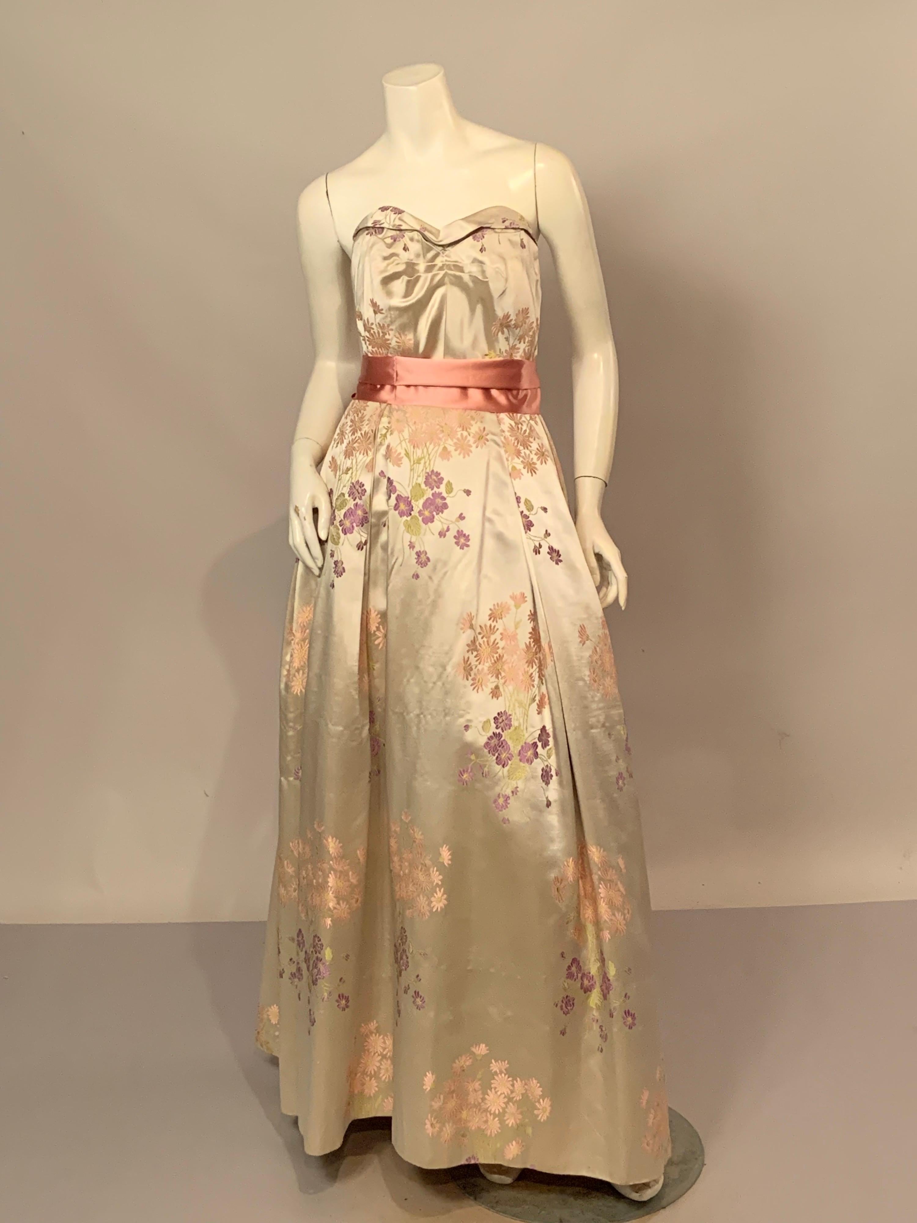 Ivory silk satin is woven with pink and green daisies and green leaves and stems, accented by a pink ribbon sash. The dress has a boned bodice with horizontal tucks at the center front and it is cut low in the back. The full skirt has pleats at the