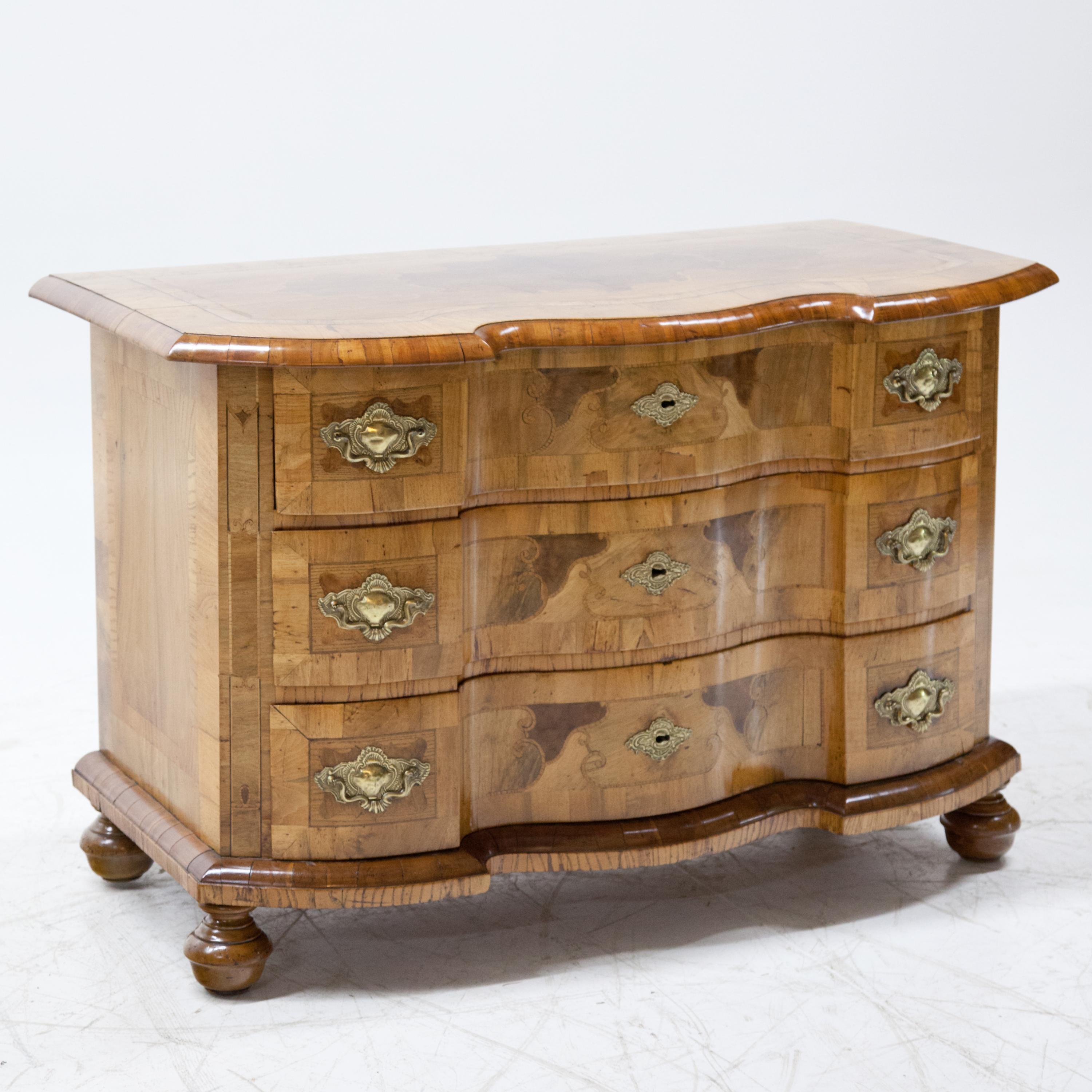 Three-drawered Baroque chest of drawers standing on spherical feet with curved front and straight sides. The top plate and the bottom cornice protrude over the sides. Walnut veneer, inlaid with C and S buckles on the drawers.
    