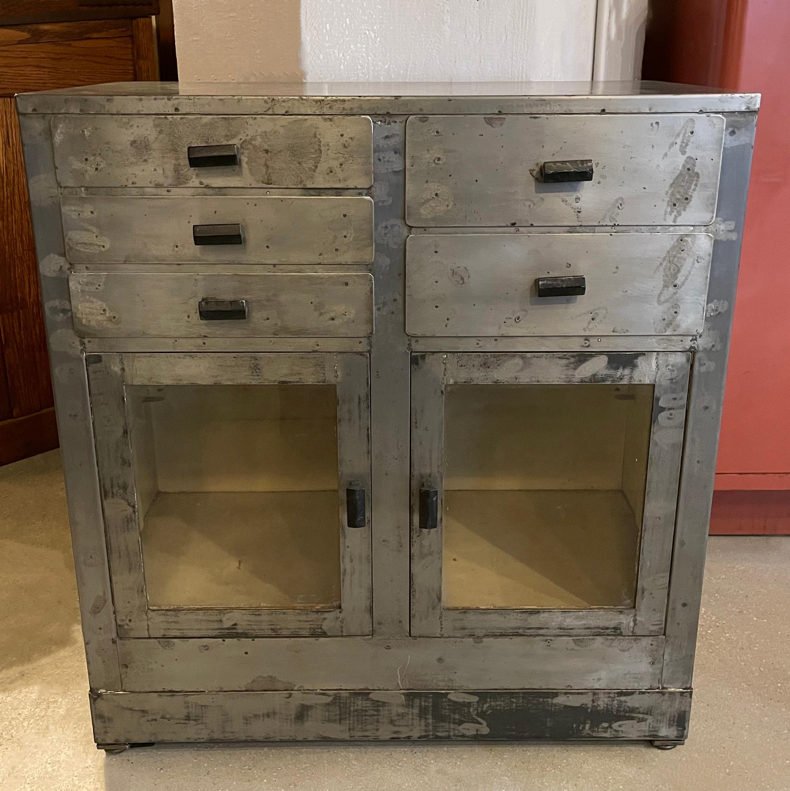 Early 20th century, industrial, deco period, brushed steel, apothecary, medicine cabinet, nurse station features top drawers and glass front bottom doors with painted interior. The black handles are Bakelite. The brushed steel exterior shows a